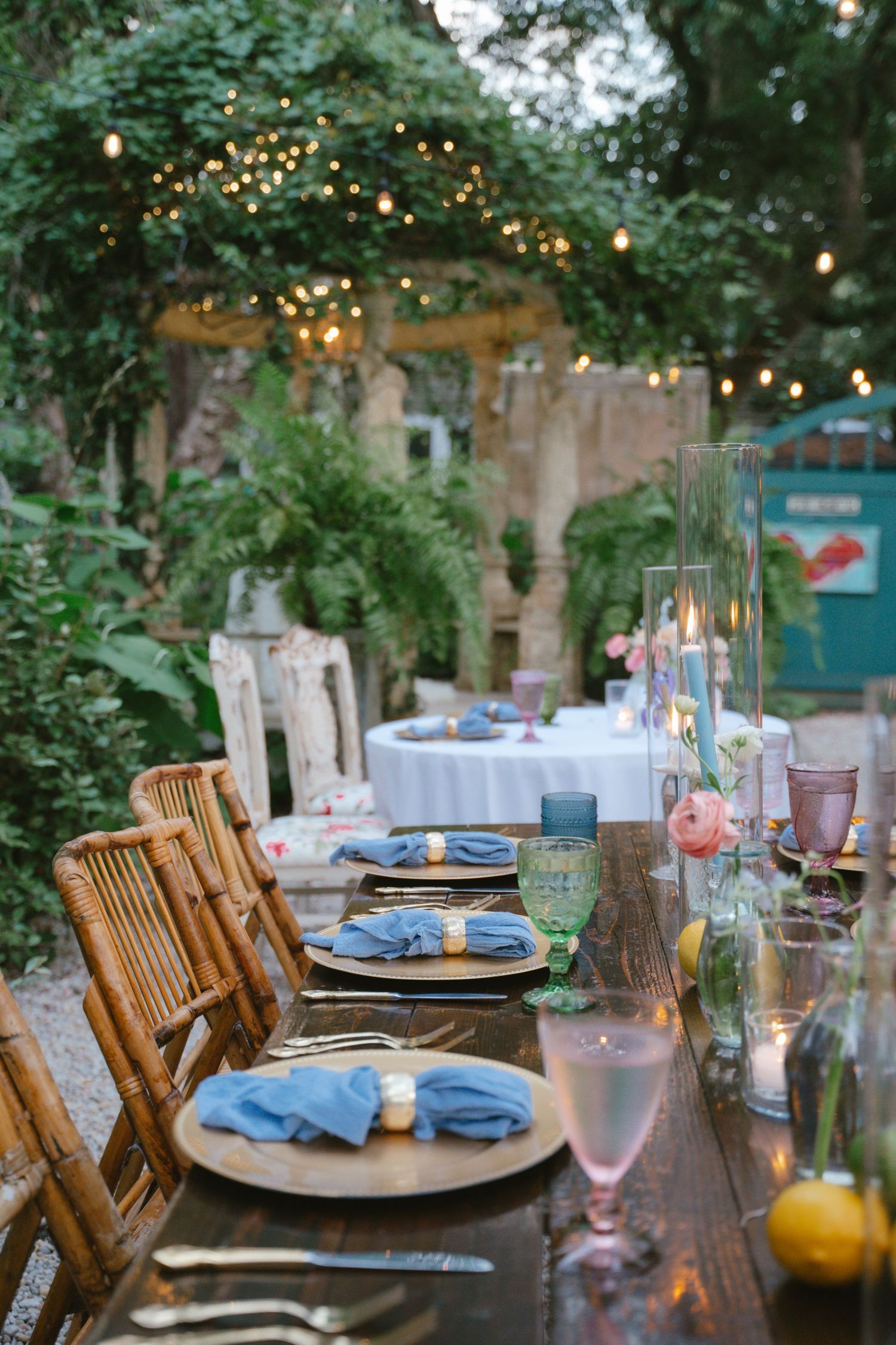 Elegant outdoor dinner table set with blue napkins, candles, and floral arrangements under string lights in a lush garden.