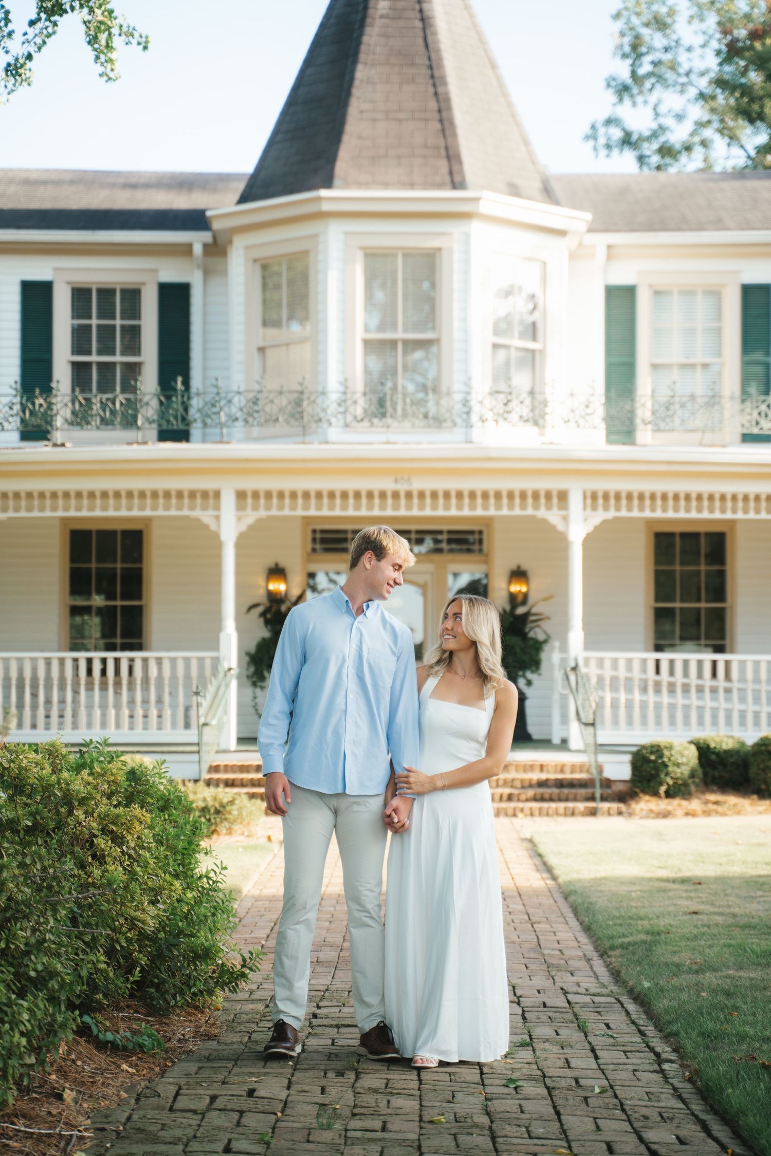 Couple holding hands smiling at each other in front of a charming white Victorian house with wrap-around porch.