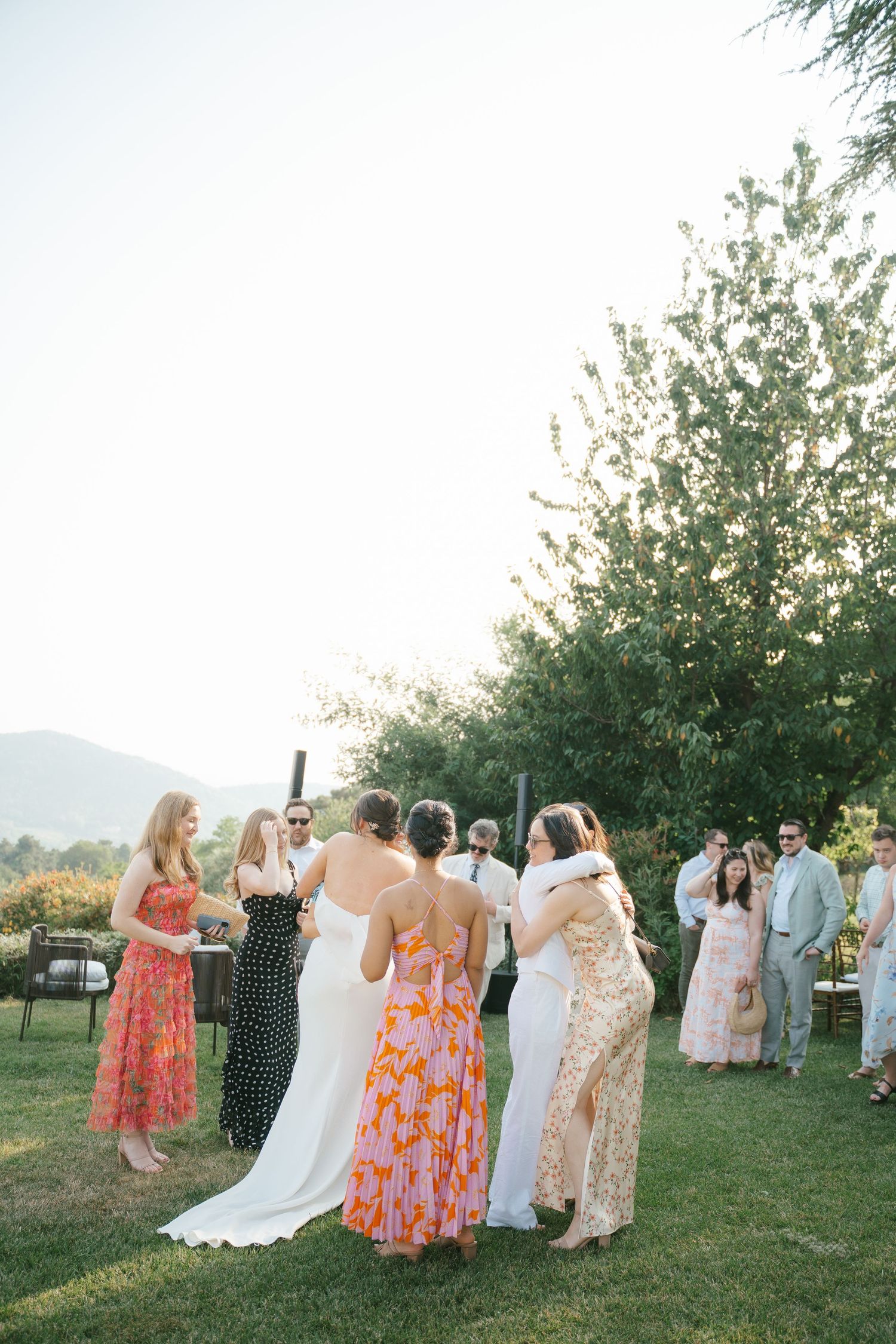 Wedding guests in colorful summer dresses gather outdoors for group photos.