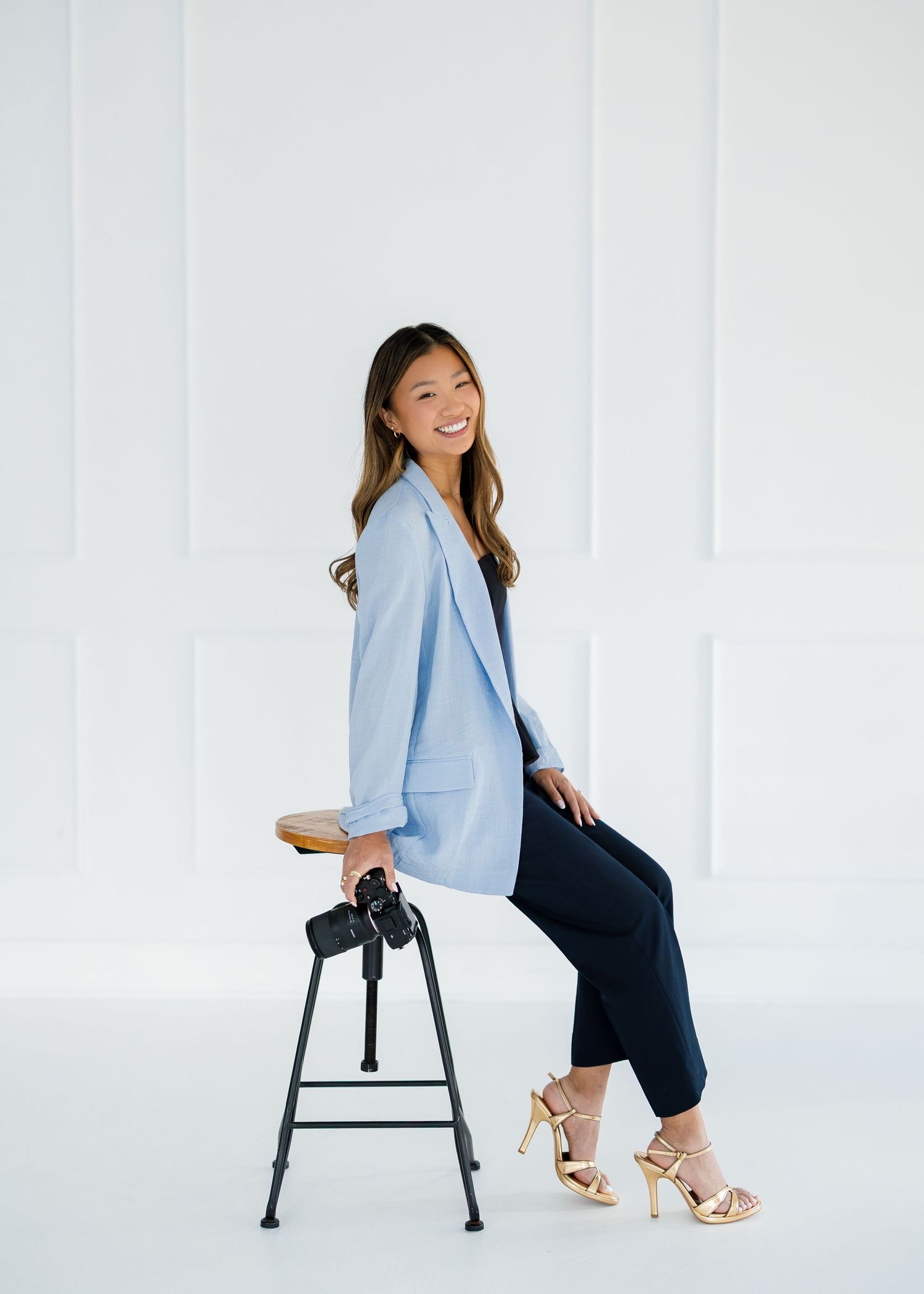 Professional in light blue blazer and dark outfit posing with camera equipment in bright studio.