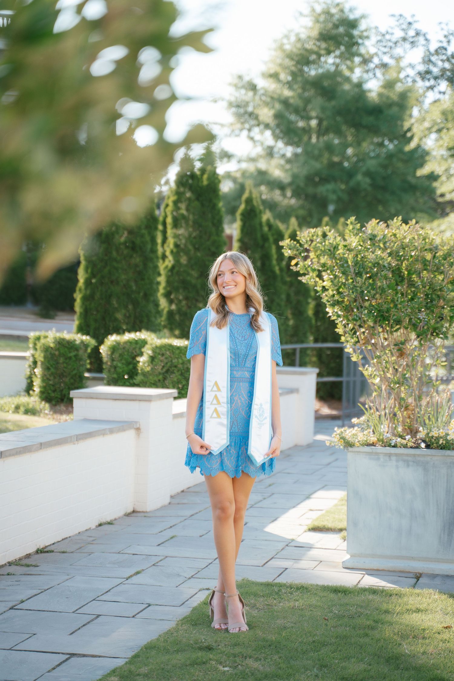 A person wearing a denim vest and white dress poses on stone steps near garden shrubs.