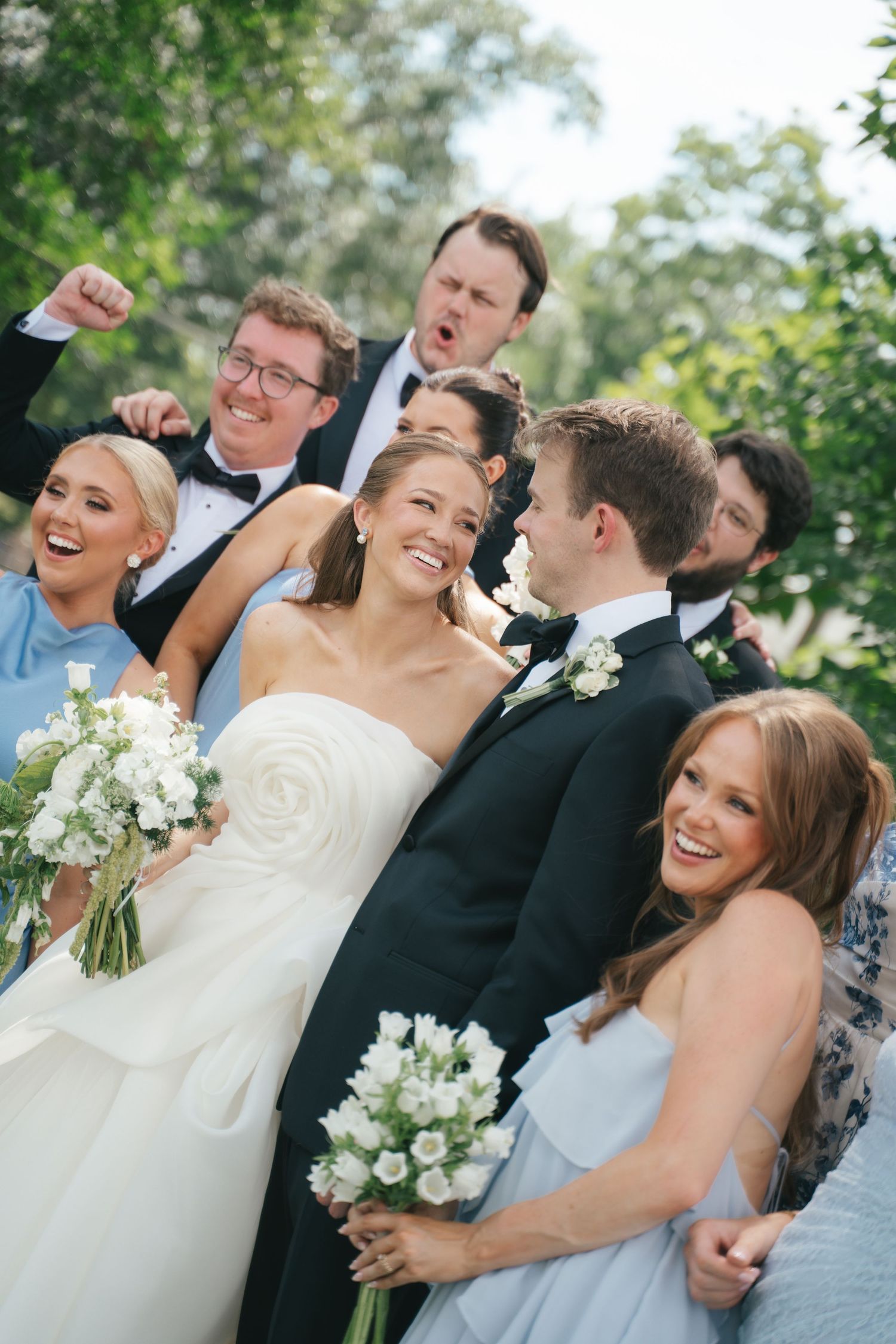 Joyful wedding party celebrating with bride and groom, bridesmaids holding white bouquets, outdoors with lush greenery.
