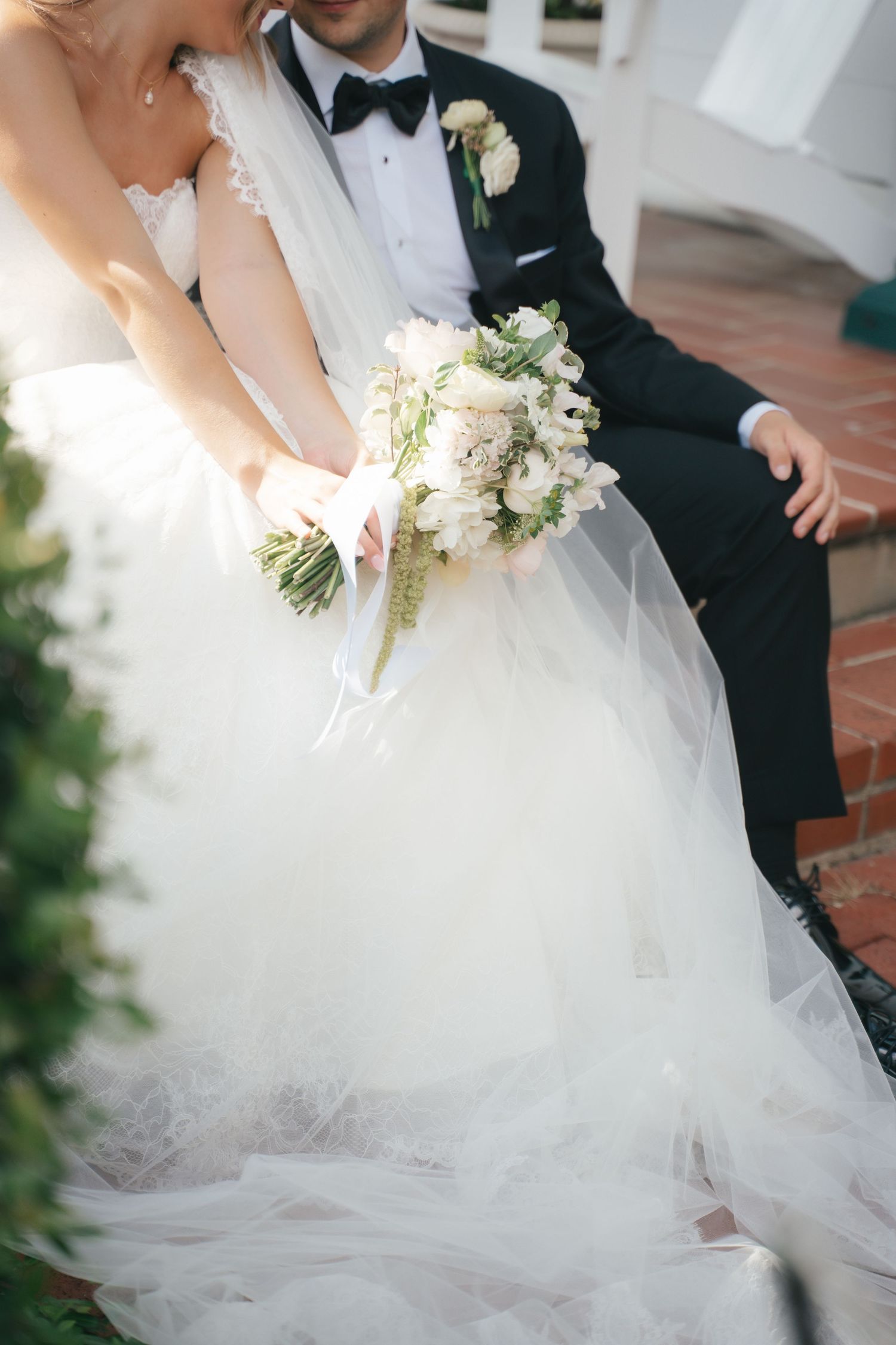 Bride in white lace ballgown holding white floral bouquet beside groom in black tuxedo on brick steps.