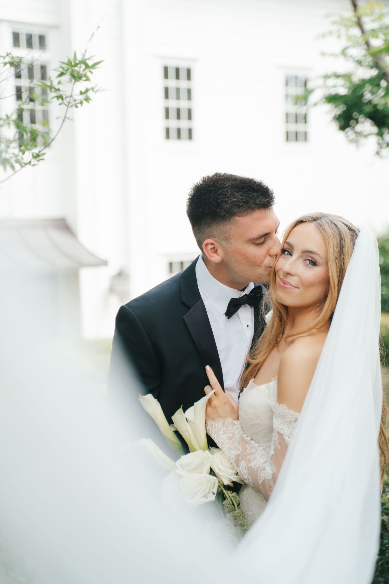 Groom in black tuxedo kissing bride's cheek as she smiles holding white bouquet outside elegant white building.