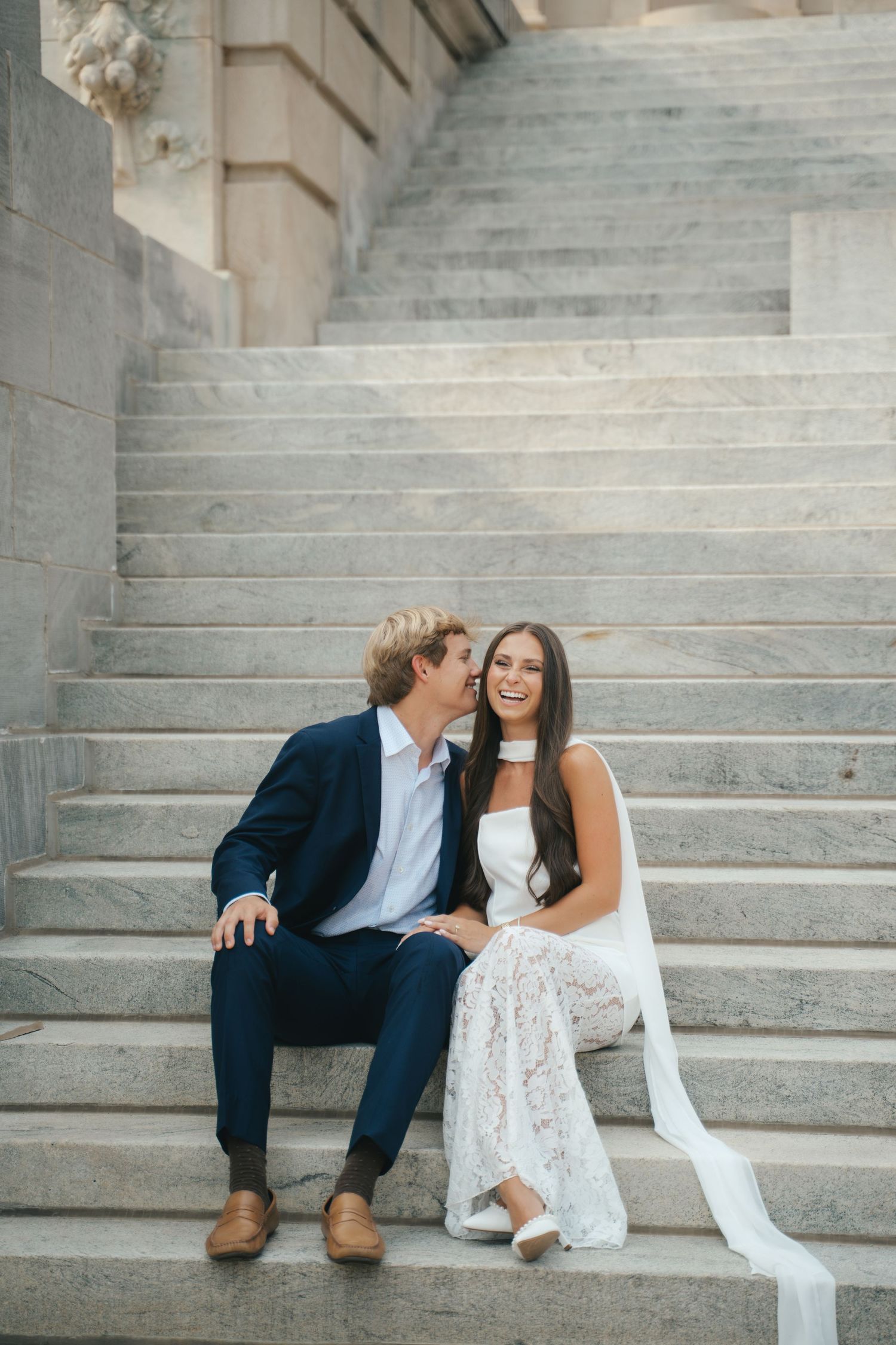A couple in wedding attire sits on marble steps, sharing a tender moment as the groom whispers to his smiling bride.