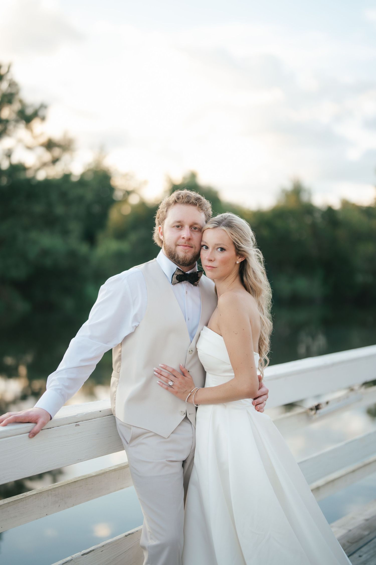 Bride and groom pose on a white bridge at sunset, with lush green trees and a reflective lake in the background.