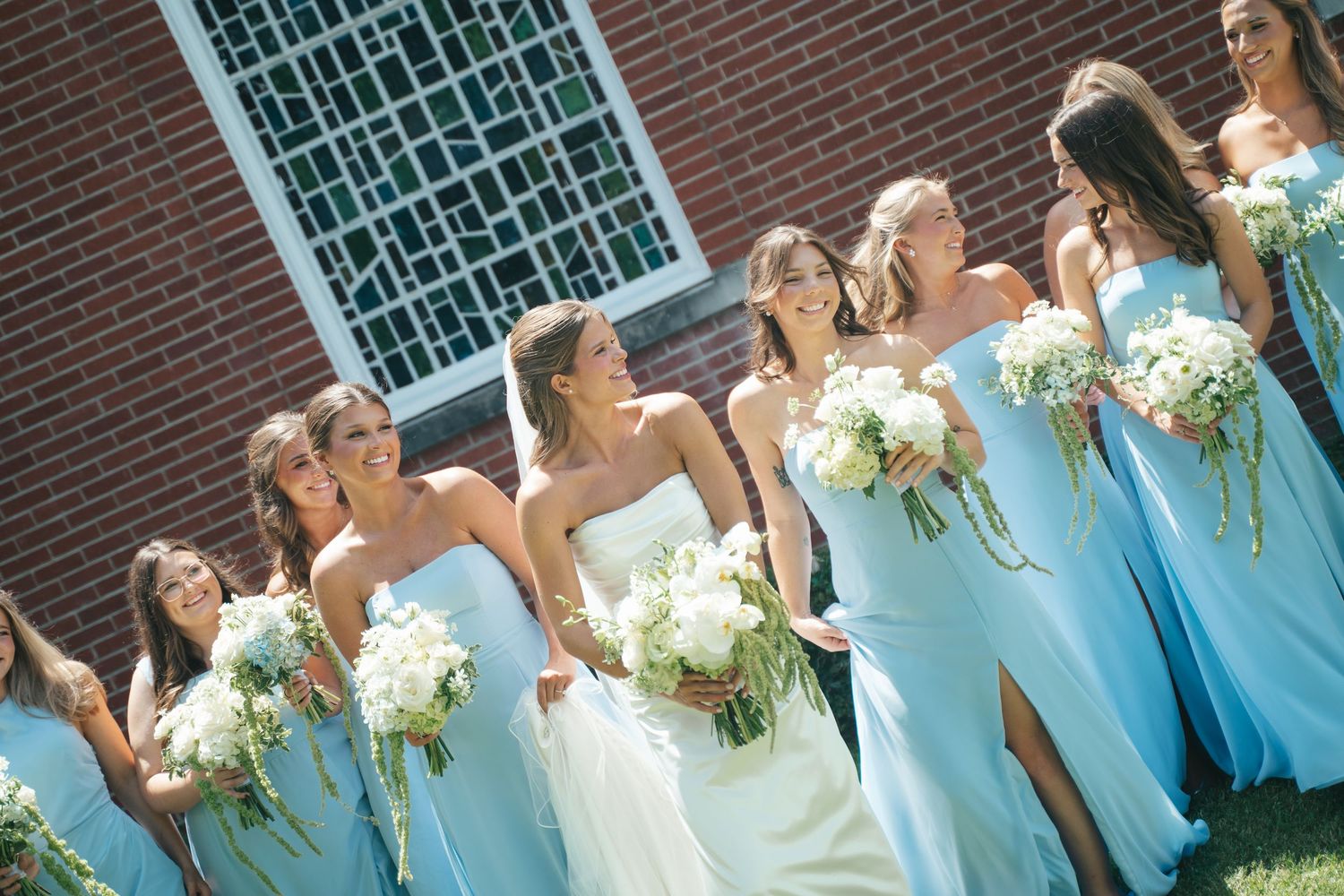Bride and bridesmaids in light blue gowns holding white bouquets pose together outdoors near a brick building.