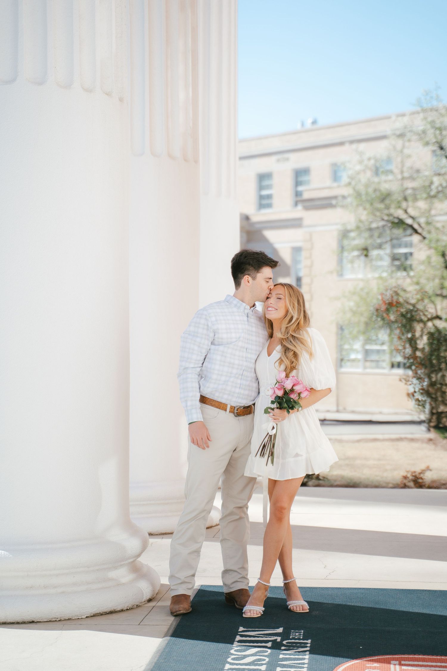 Couple shares intimate moment between white columns on sunny day.