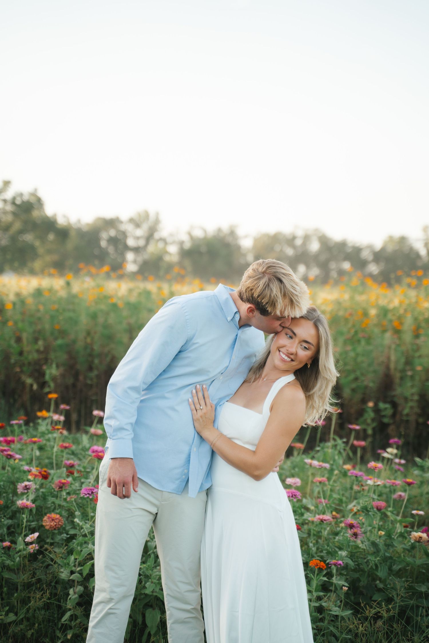 A couple embraces in a vibrant flower field, the man kissing her cheek as she smiles in a white dress.