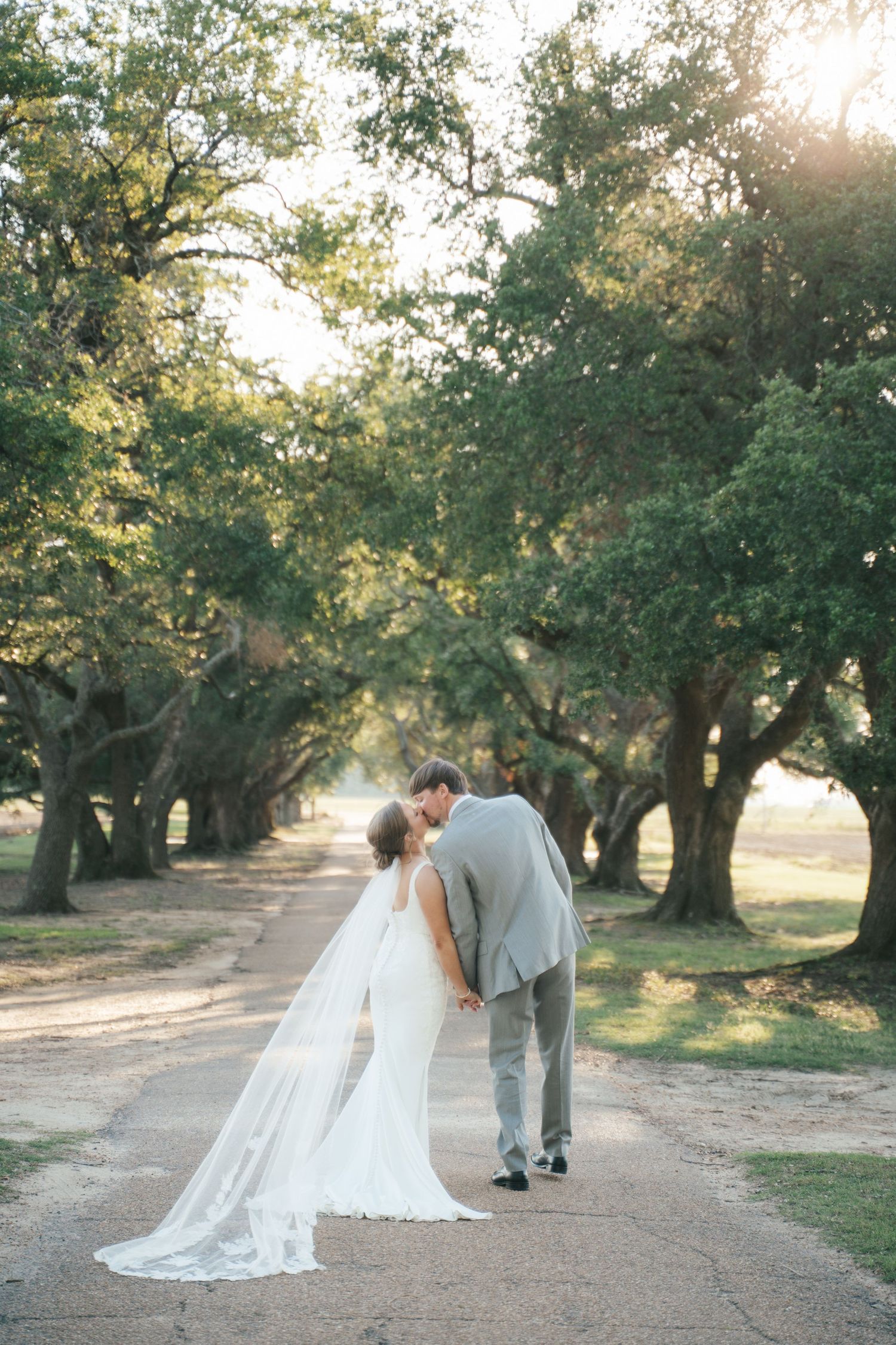 Bride and groom share a romantic kiss on a tree-lined path, bride wearing a flowing white gown with long veil.