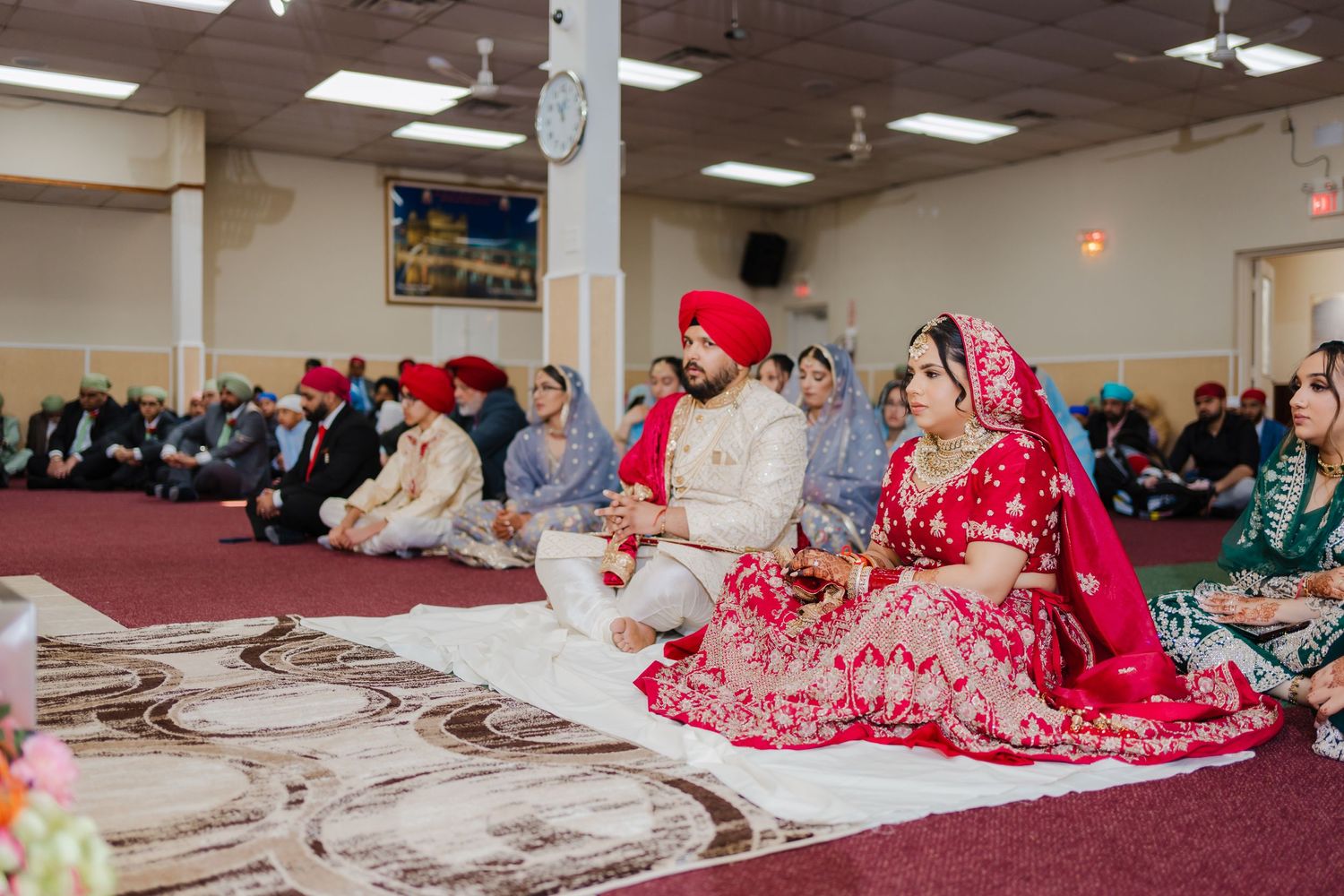 Traditional Sikh wedding ceremony taking place in a temple with guests seated on the carpeted floor during prayer.