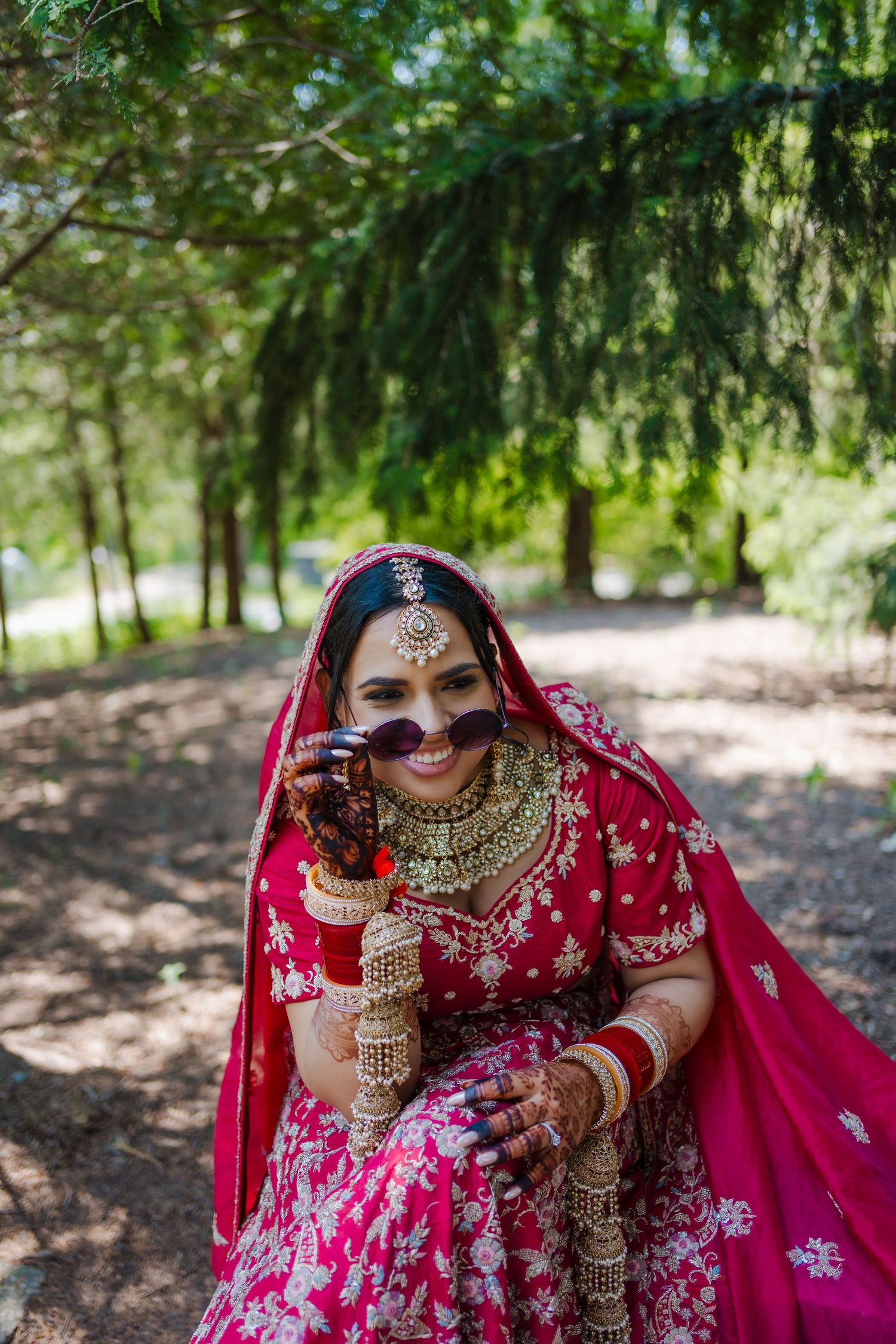 Traditional bride in red embroidered lehenga and gold jewelry poses elegantly under evergreen trees in outdoor setting.