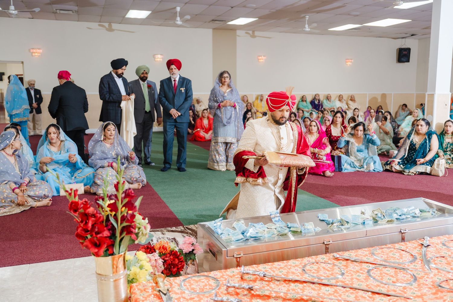 Traditional Sikh wedding ceremony in a Gurdwara with guests seated on floor and decorative floral arrangements.