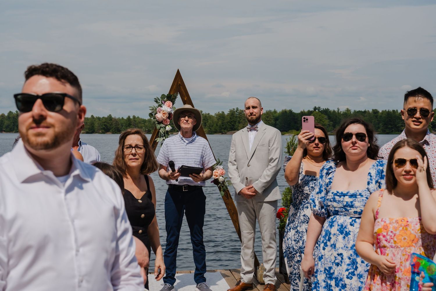 A group of wedding guests standing together by a waterfront on a cloudy day.