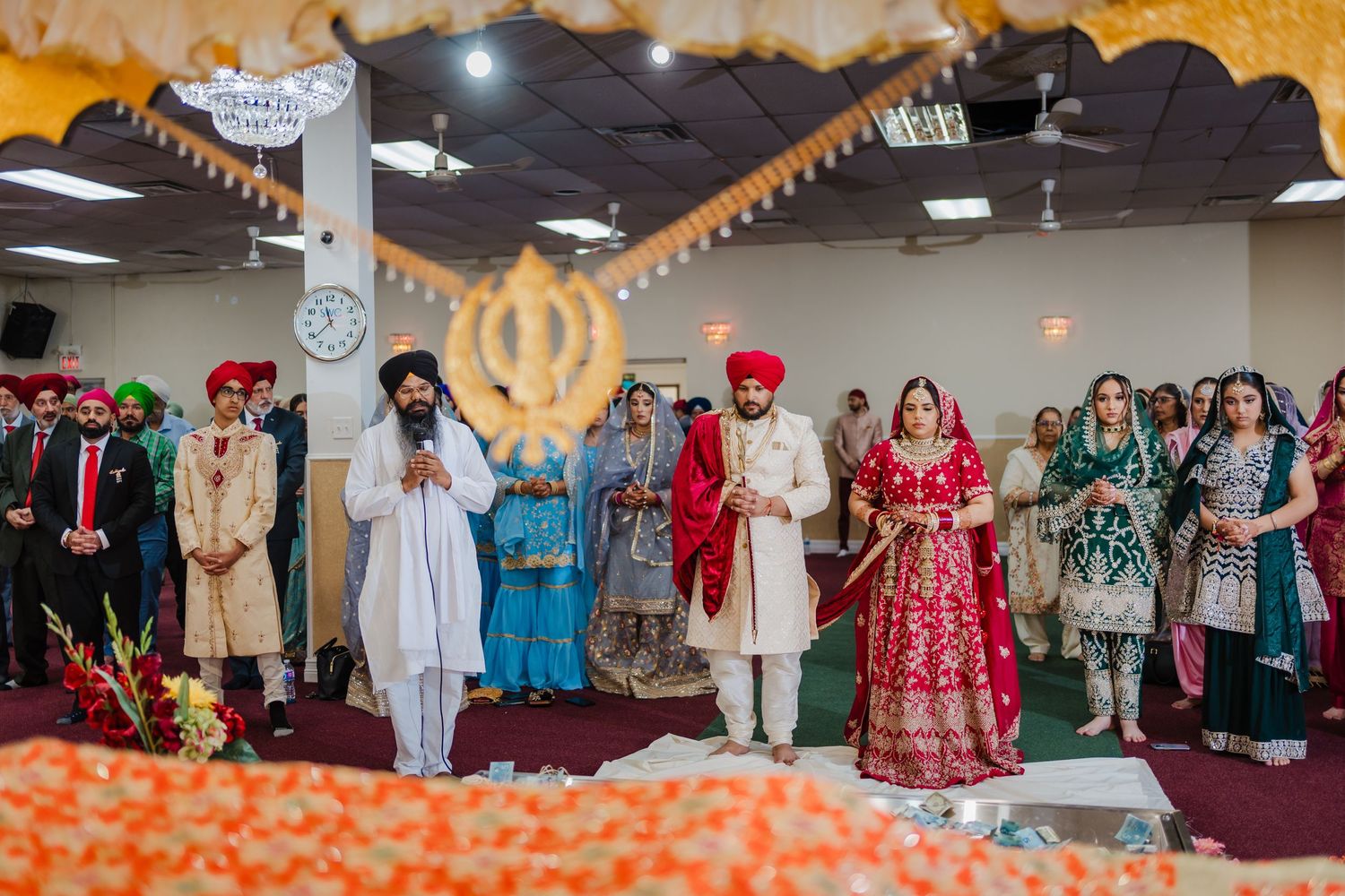 Traditional Sikh wedding ceremony taking place indoors with guests in colorful traditional attire gathered around.