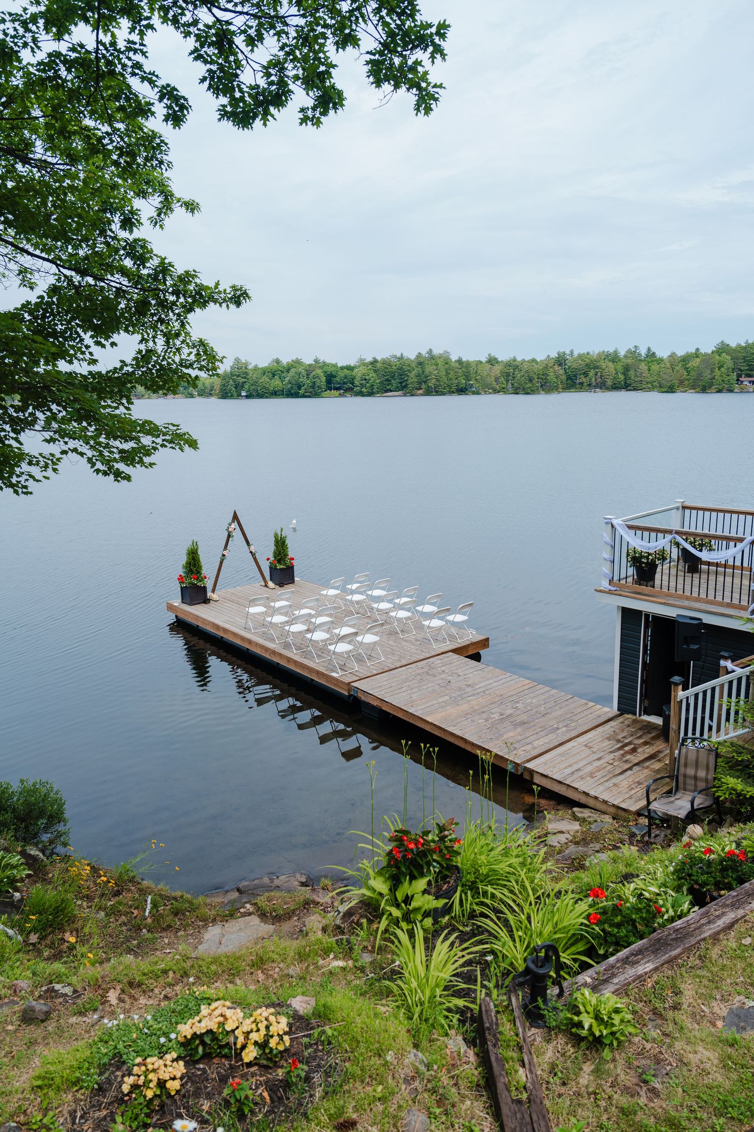 Wooden dock with glassware setup on a serene lake surrounded by trees and landscaped garden on a cloudy day.