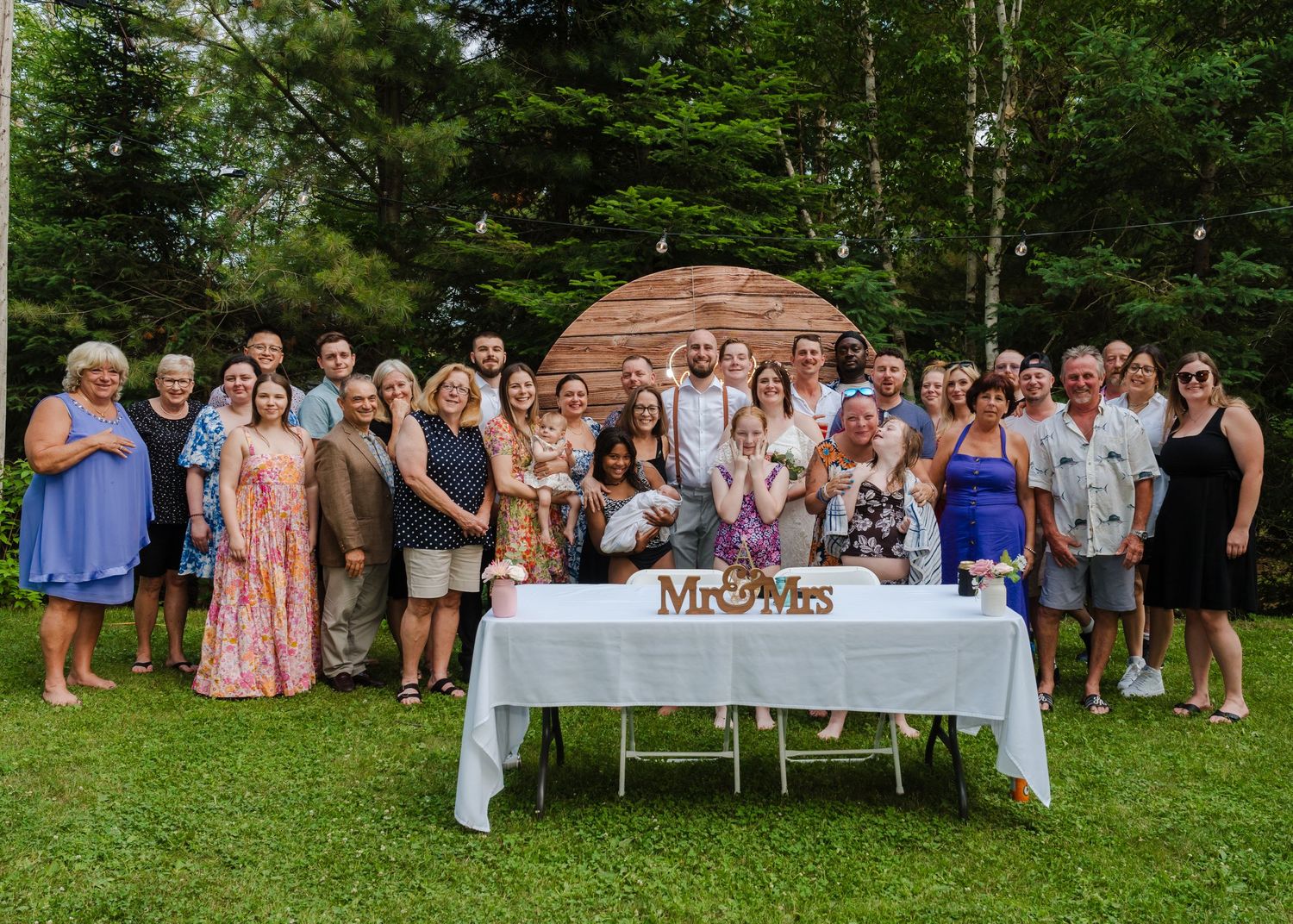 Large group gathered outdoors in front of a wooden arch structure with a white table for a special celebration.