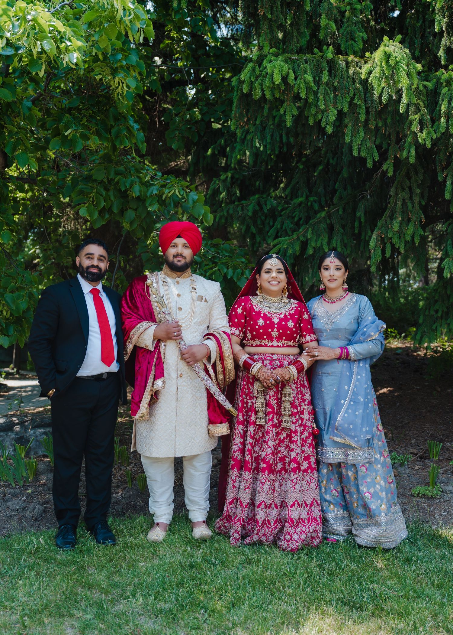 Four people in traditional Indian wedding attire pose together outdoors with greenery in the background.