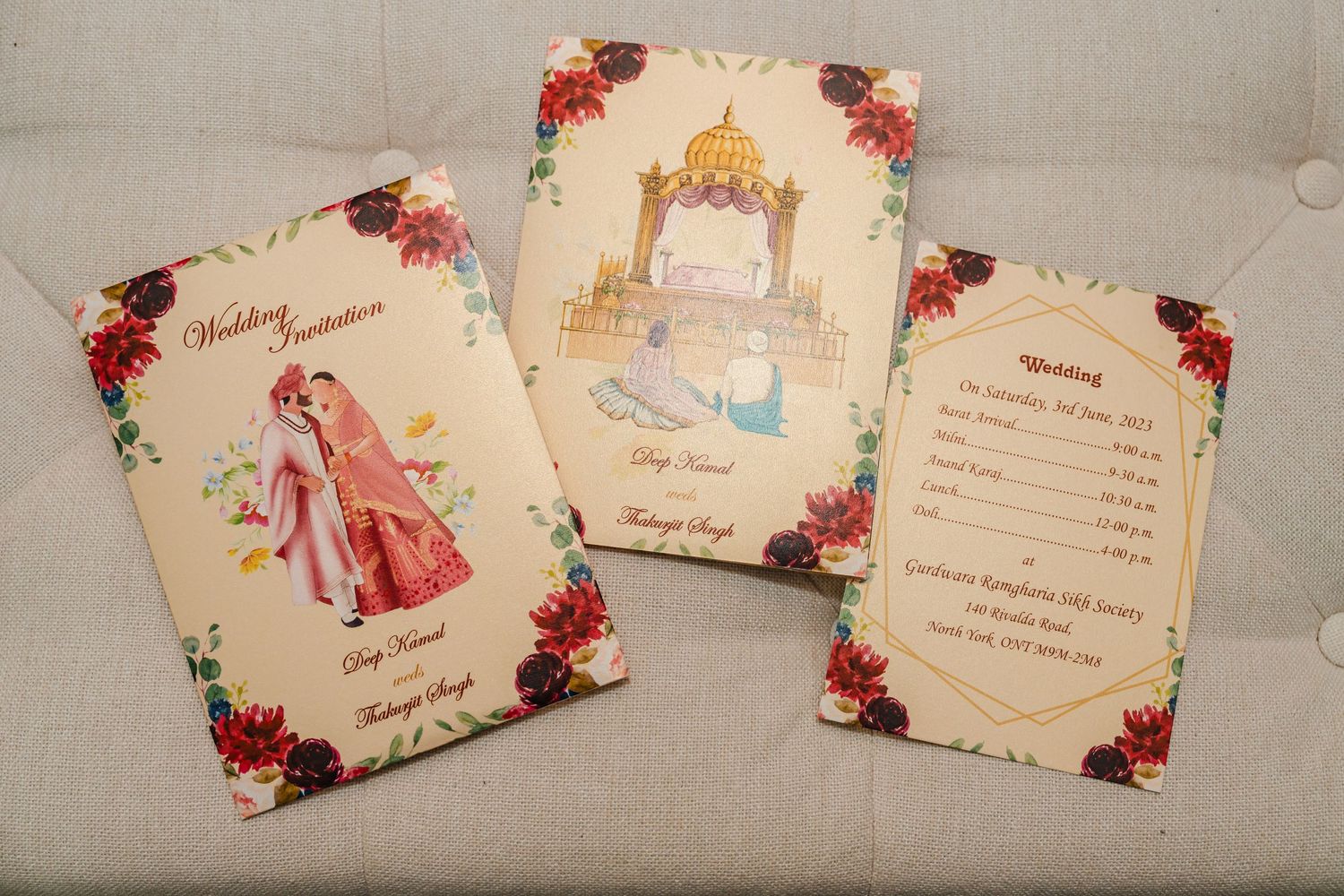 Three elegant wedding invitation cards decorated with red roses and gold designs on a light grey couch.