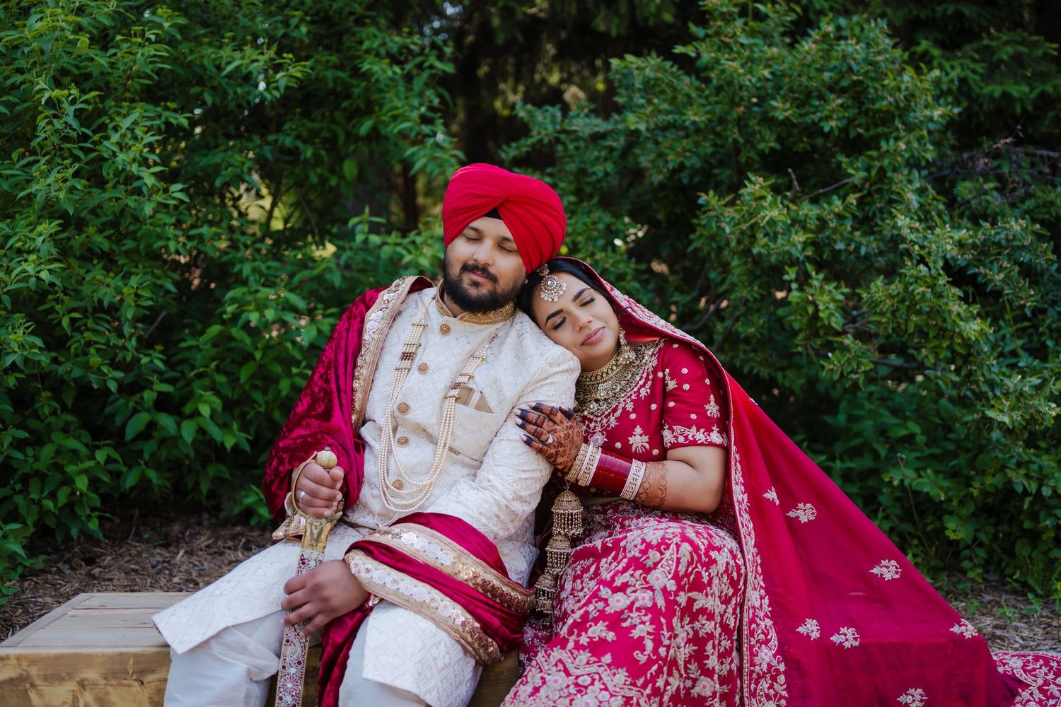 Couple in traditional red and white Indian wedding attire sitting together on outdoor stone bench.