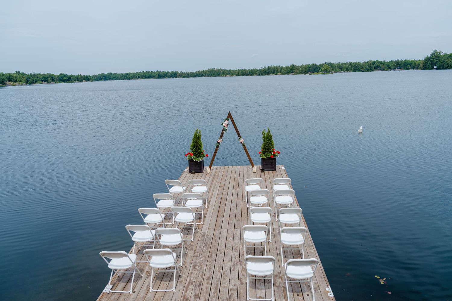 Aerial view of dock wedding setup with white chairs facing triangular arch over water.