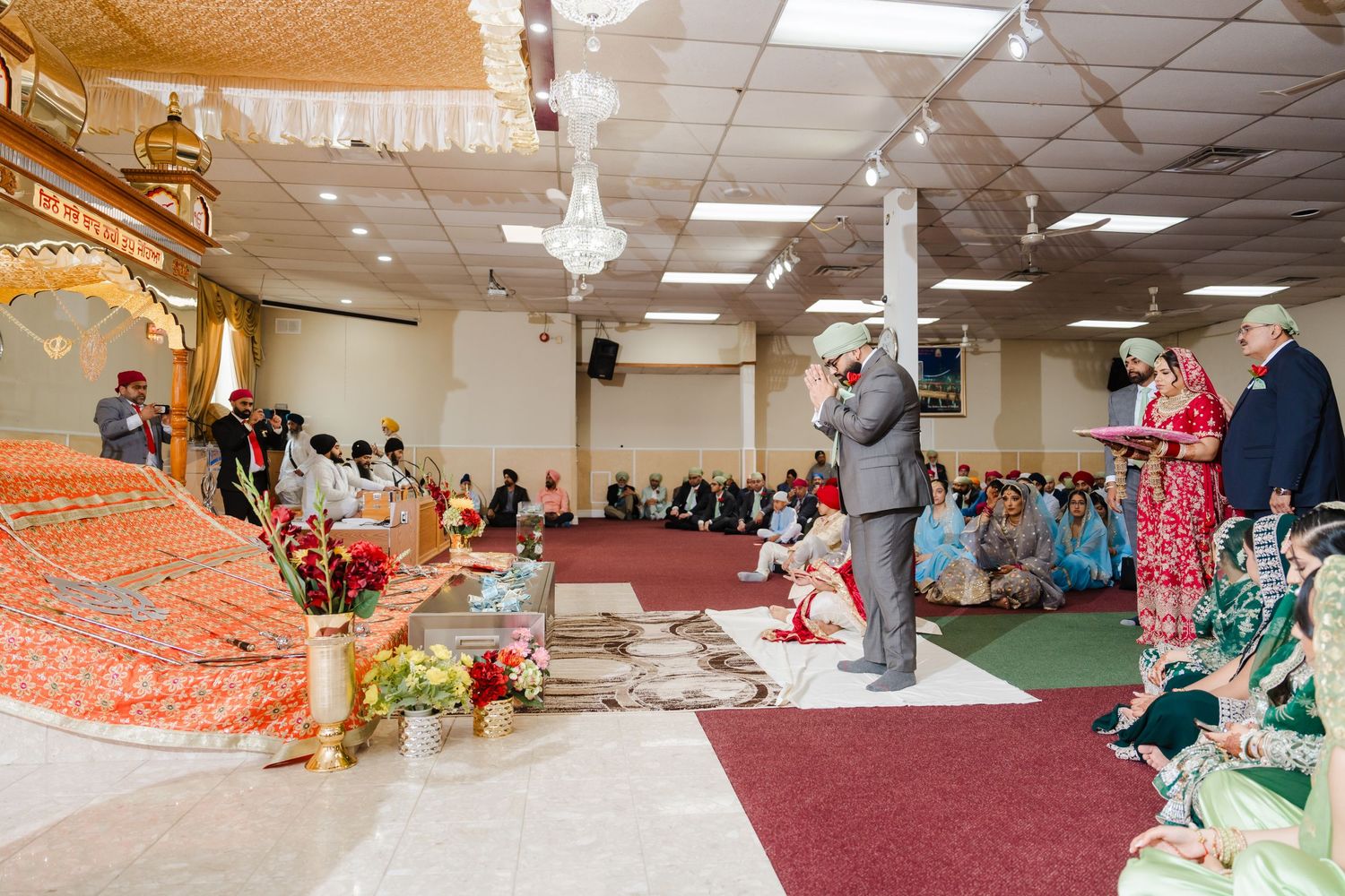 Wedding guests gather in a carpeted reception hall with decorative orange and red traditional cultural decor.