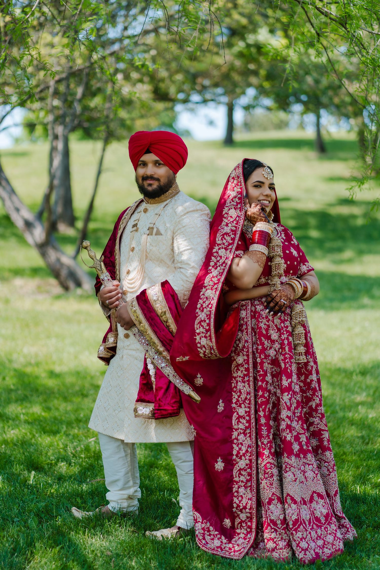A couple in traditional Sikh wedding attire poses in a garden setting with red and white cultural dress.