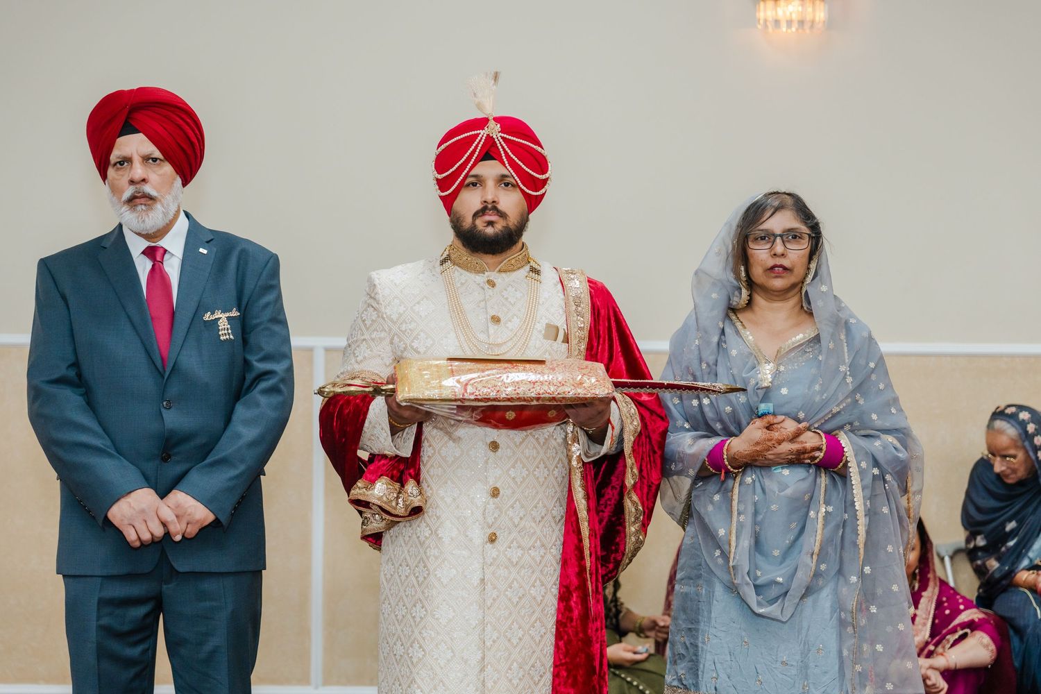 Three people in traditional Sikh wedding attire standing together during a ceremony, with red turbans and formal clothing.