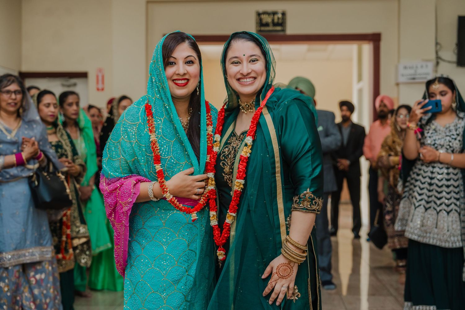 Two women wearing vibrant turquoise traditional South Asian outfits smile together at an indoor cultural event.