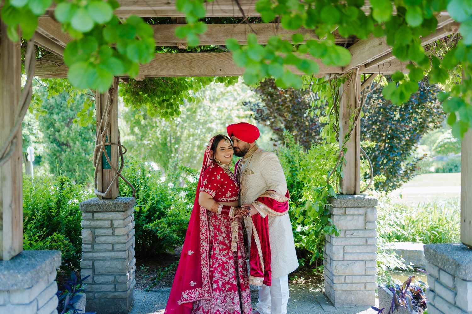A couple in traditional South Asian wedding attire embrace under a garden pergola with green vines.