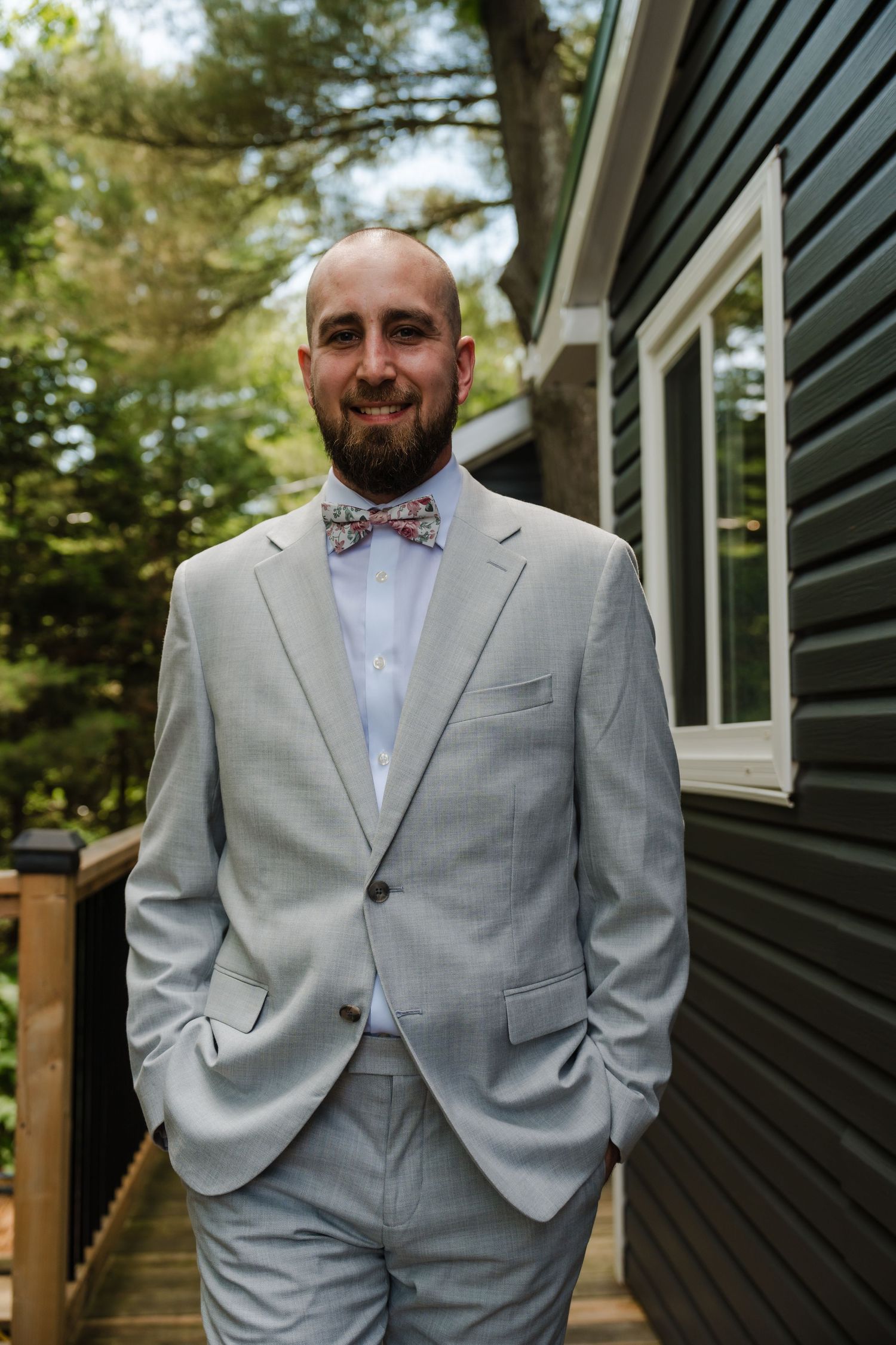 Groom in light grey suit and floral bow tie poses confidently on deck of dark building.