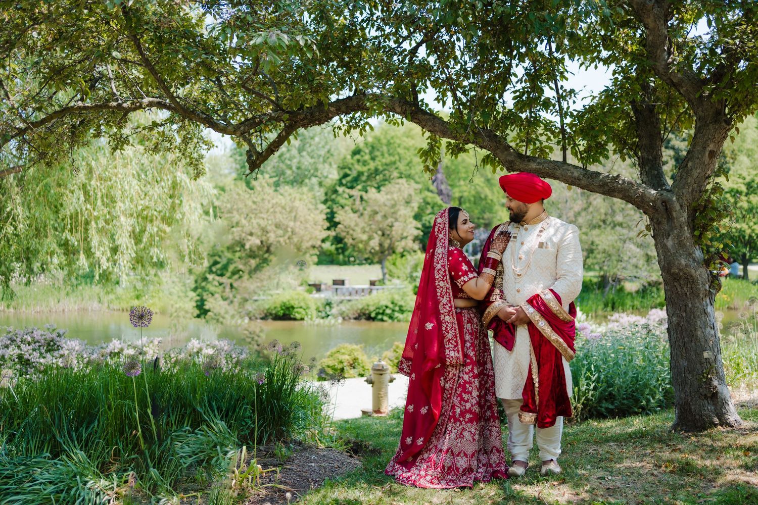 A couple in traditional Punjabi wedding attire stands under a willow tree in a garden setting.