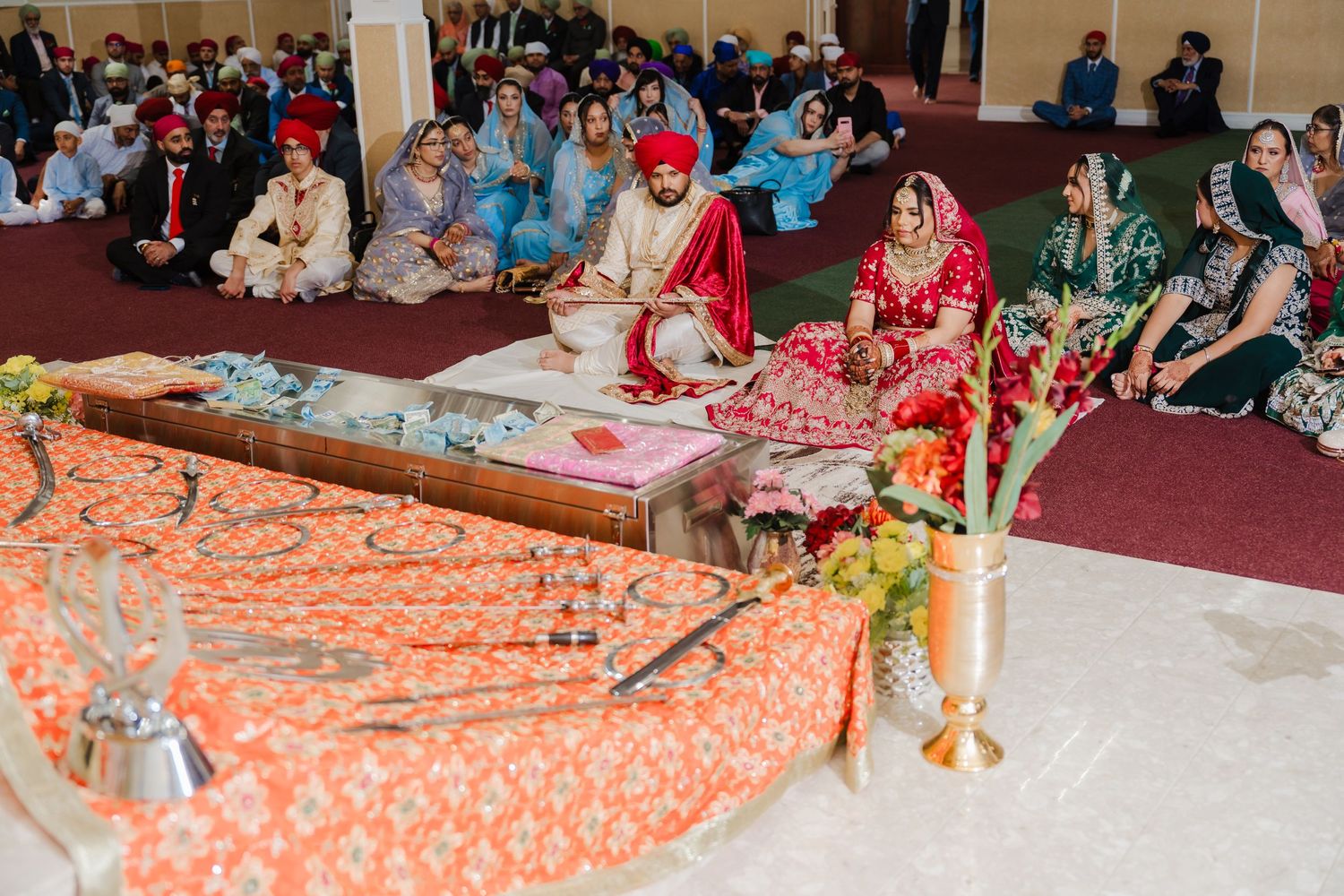 Traditional Indian wedding ceremony gathering with guests seated on floor around decorative orange and red textiles.