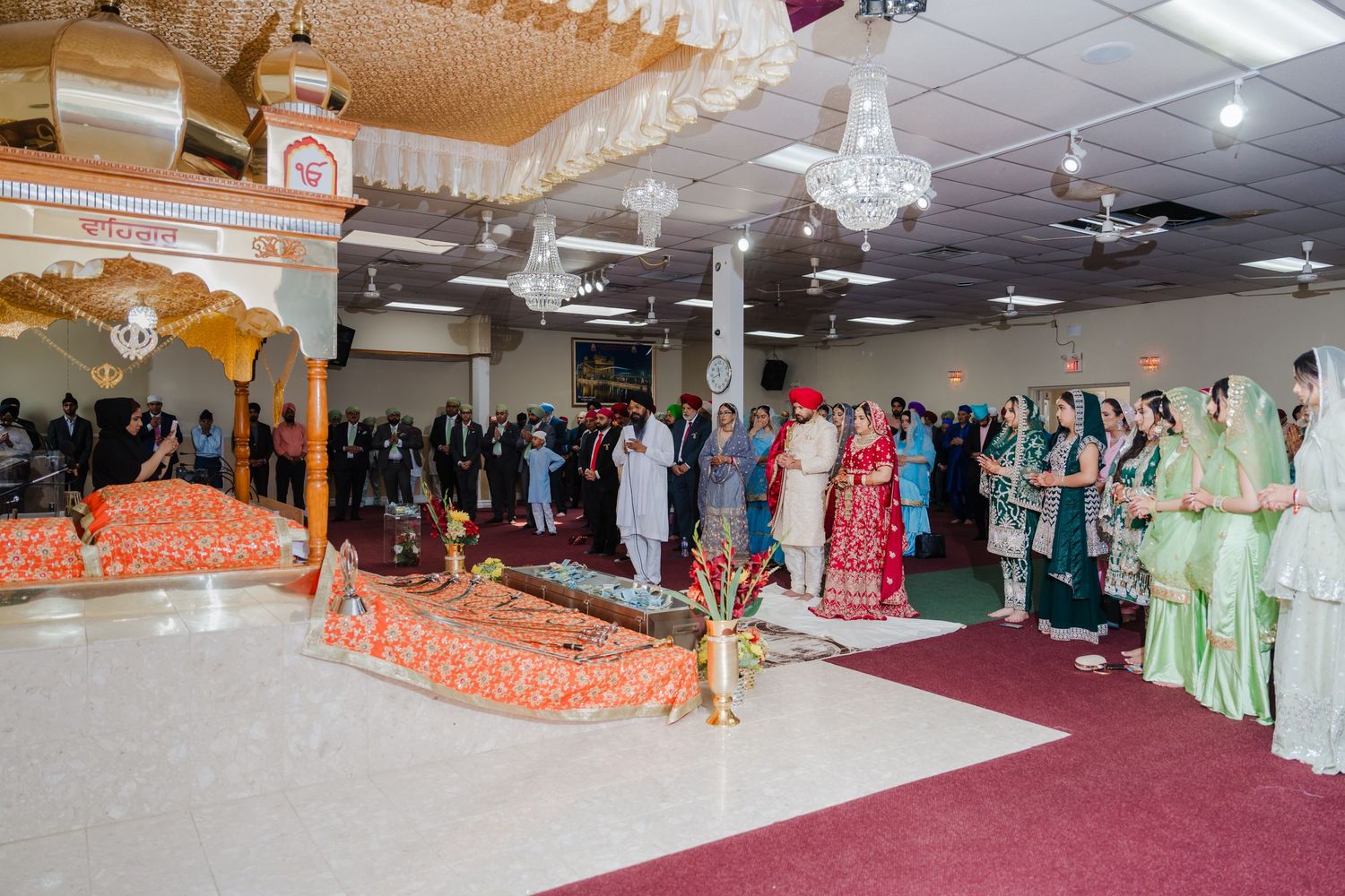 Guests in vibrant traditional clothing stand in rows during a cultural celebration in a decorated indoor venue.