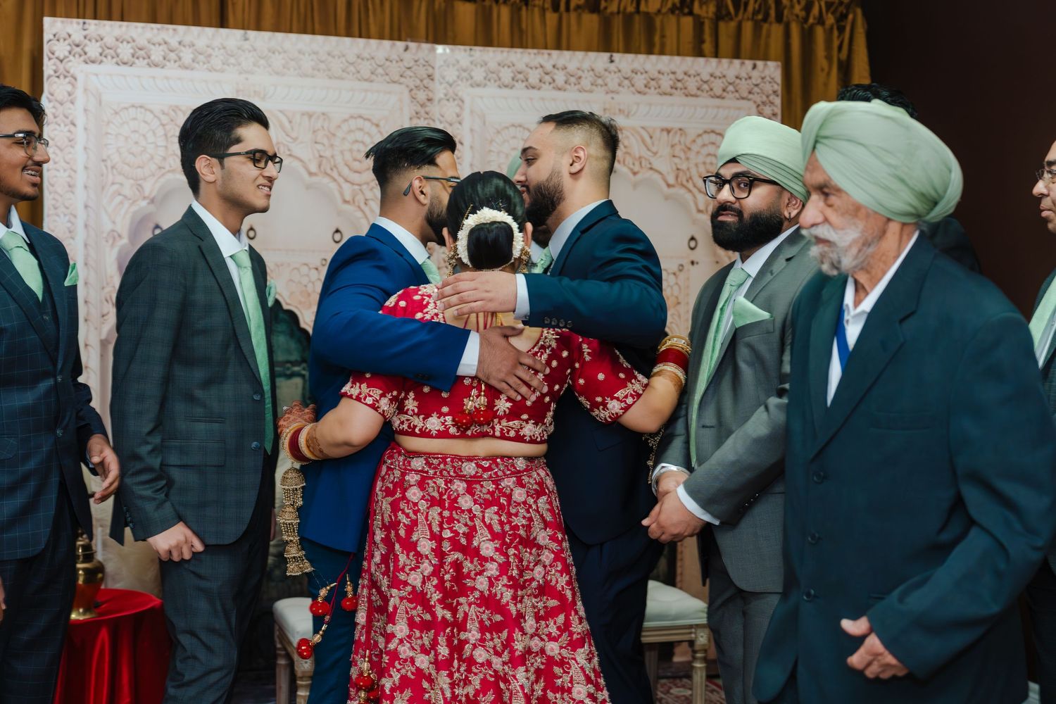 A couple in traditional attire embrace during a wedding celebration while guests in suits and turbans look on.