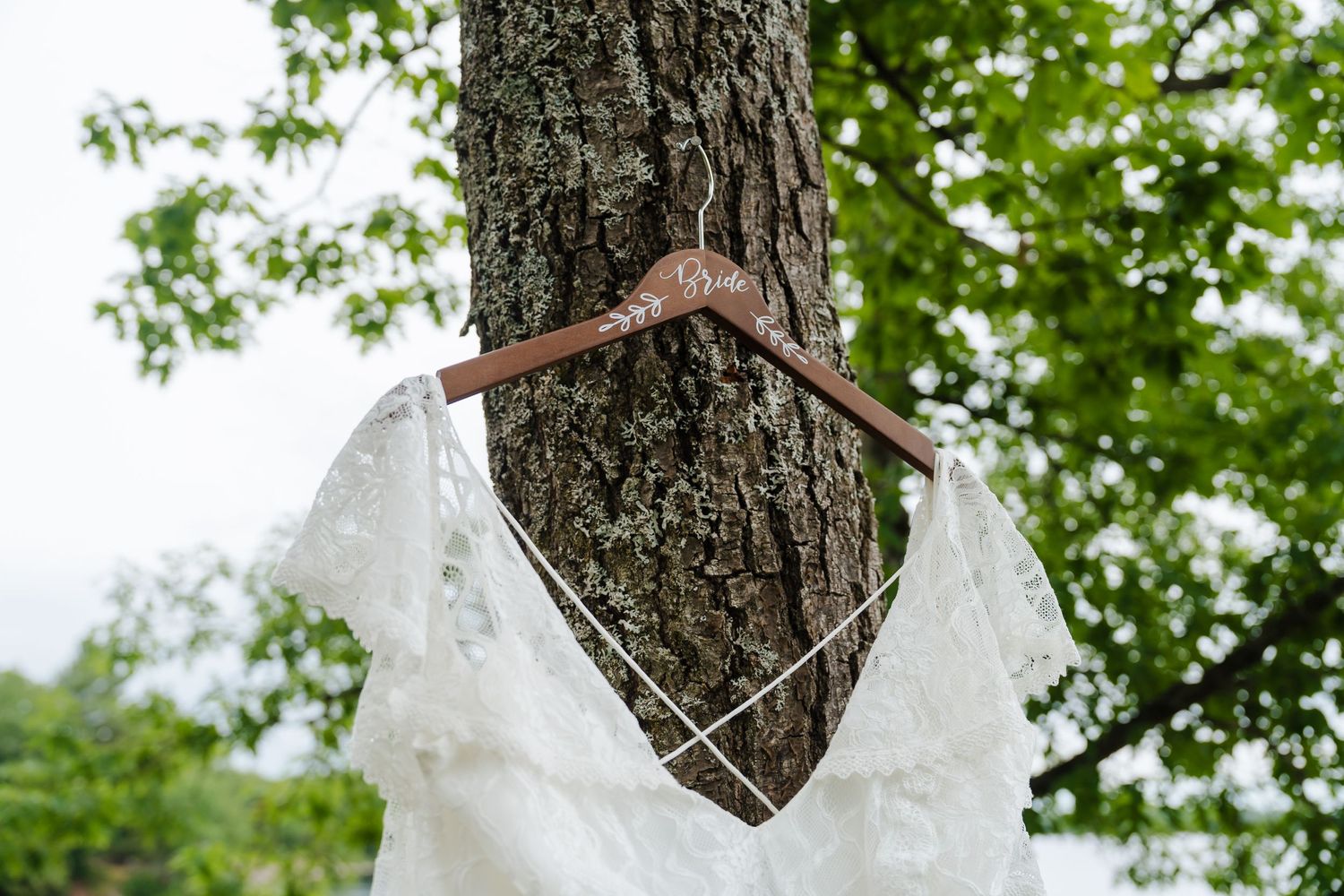 A delicate white lace wedding dress hangs on a wooden hanger against a tree trunk with green foliage in the background.