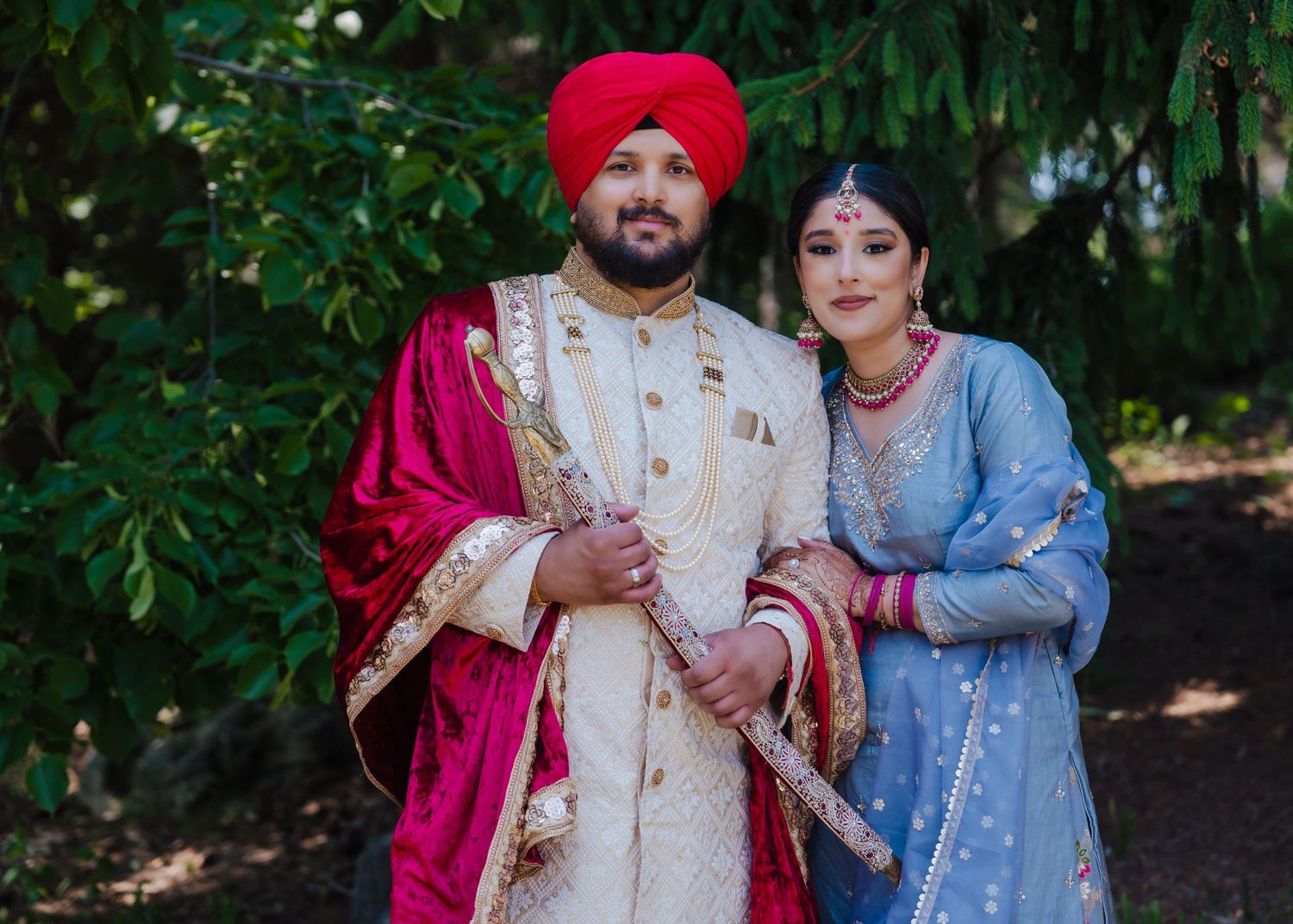 A couple dressed in traditional South Asian wedding attire, with vibrant red and light blue outfits, pose outdoors.