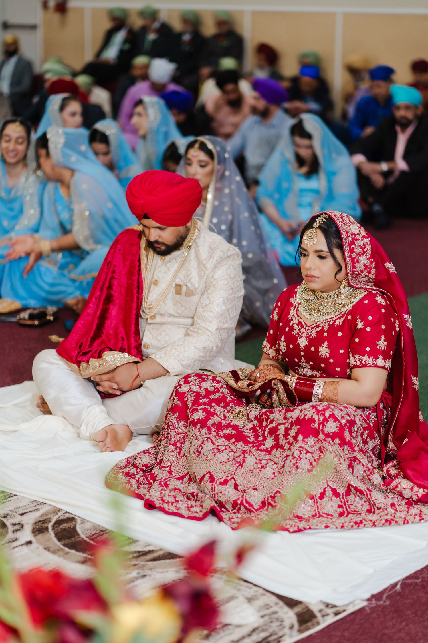 Bride and groom in traditional Sikh wedding attire sit together during ceremony with guests in background.