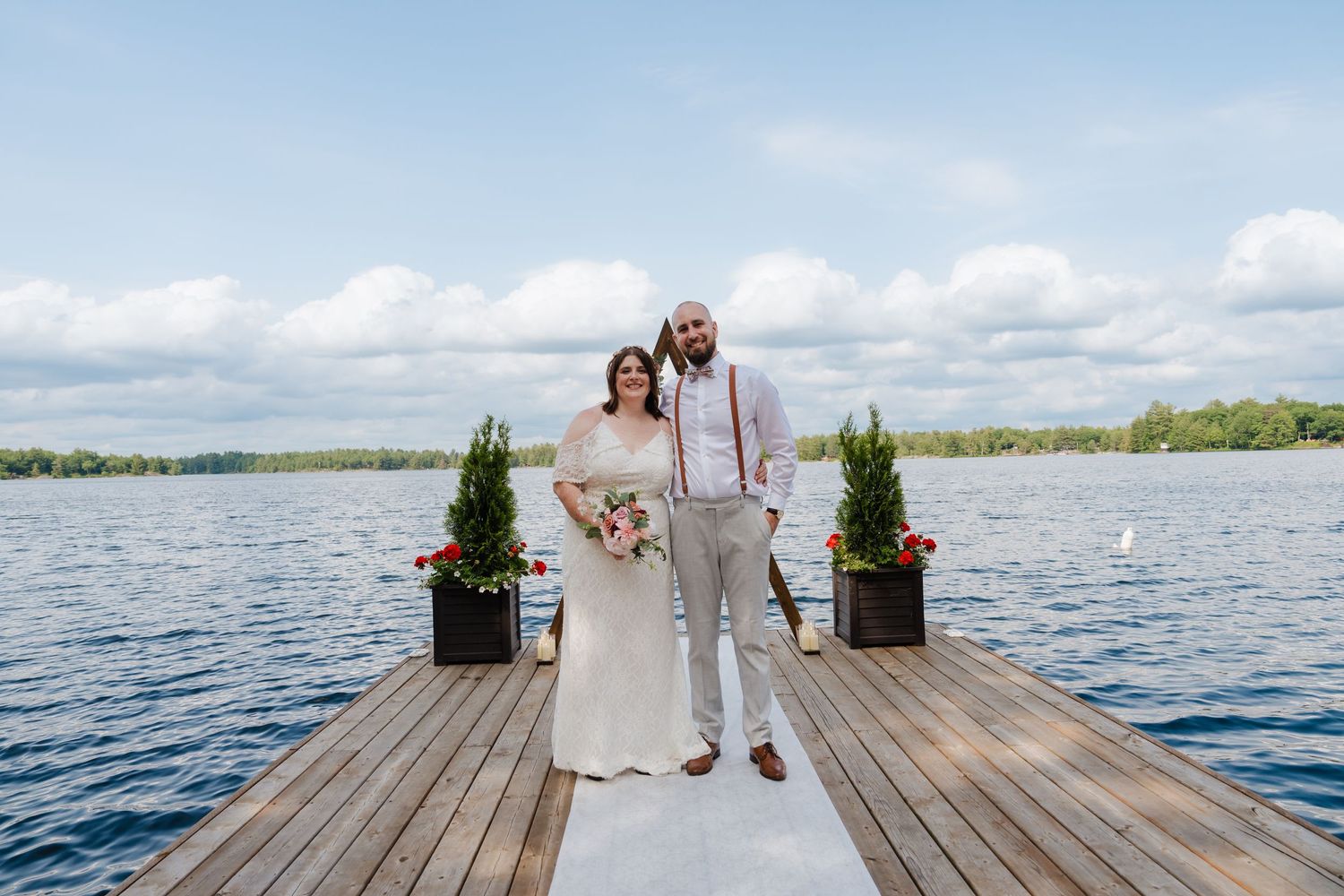 A couple stands on a wooden dock overlooking a lake with potted evergreens and white clouds in the sky.