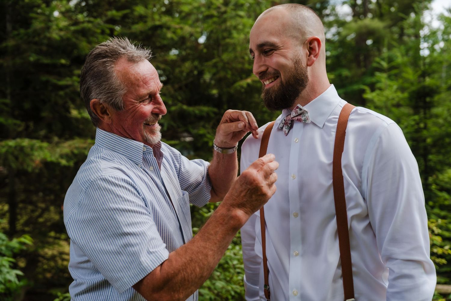 Two men share a special moment while adjusting formal attire with suspenders during wedding preparations.