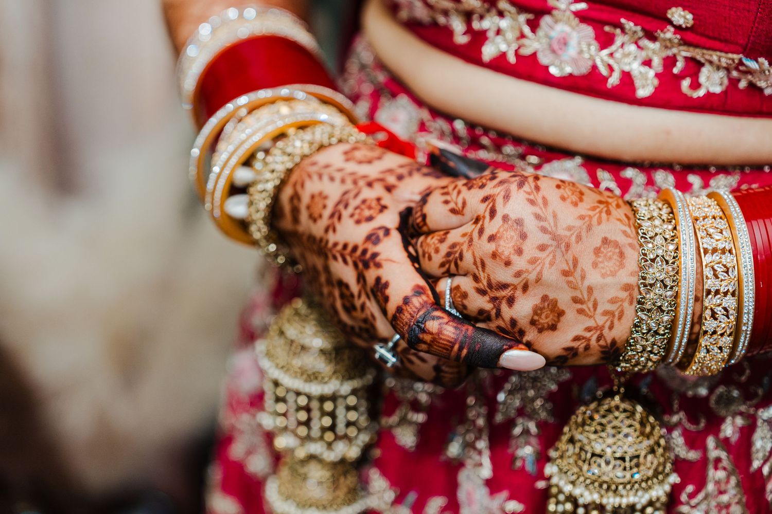 Intricate henna designs and traditional gold bangles adorn hands wearing red bridal attire at an Indian wedding ceremony.