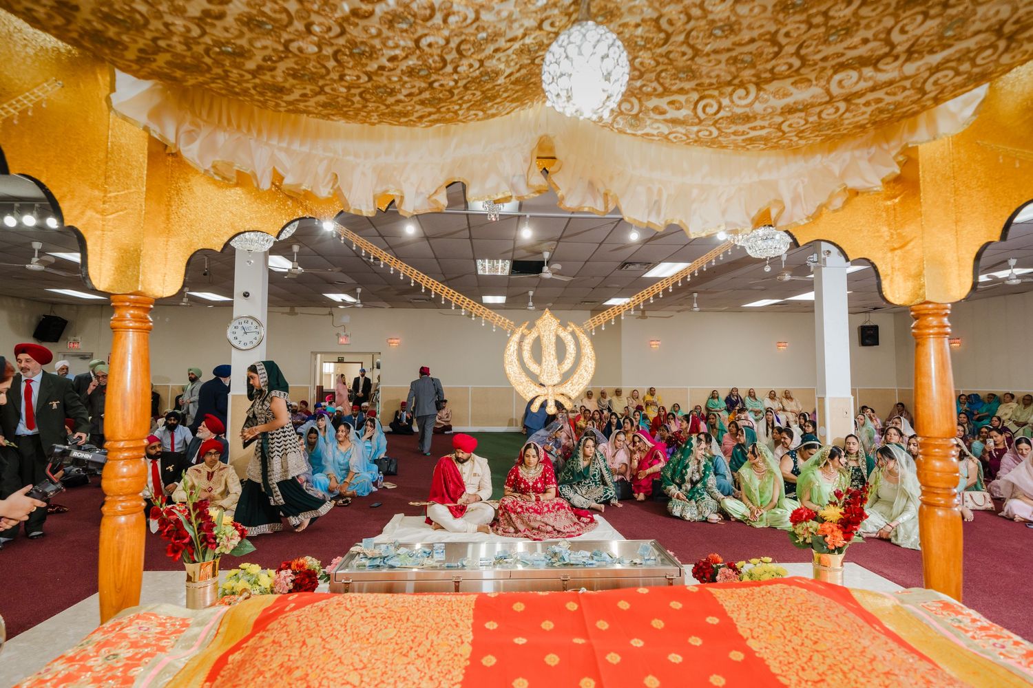 Interior of ornately decorated Gurdwara temple with golden arches and crowd gathered for Sikh ceremony.