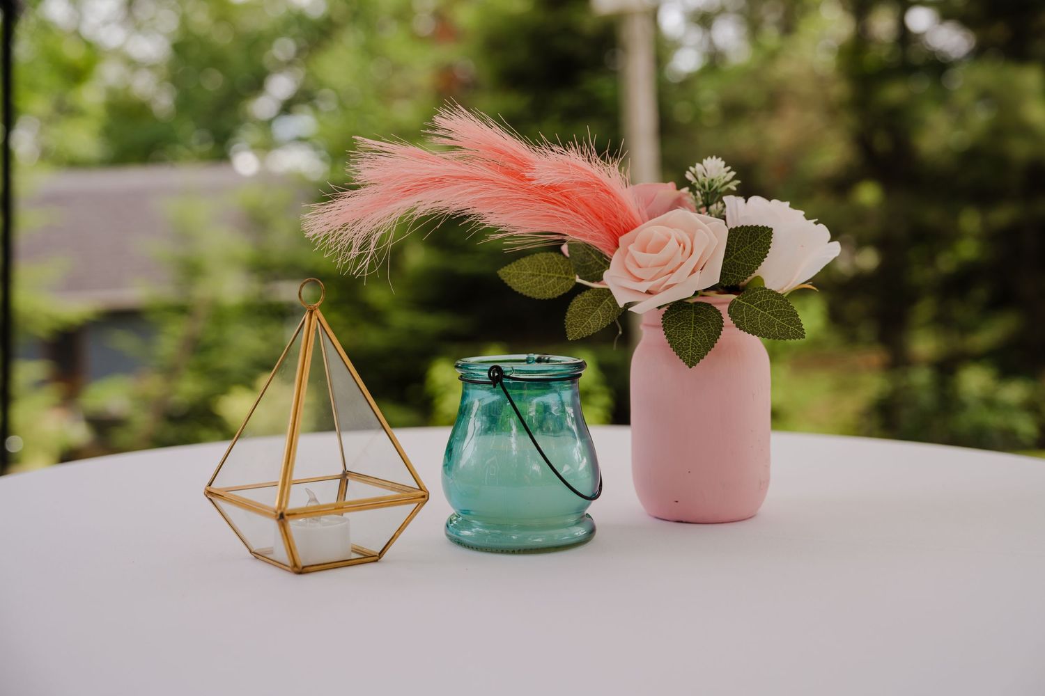 Decorative geometric gold terrarium, blue glass jar, and pink vase with feathers and flowers on white table outdoors.