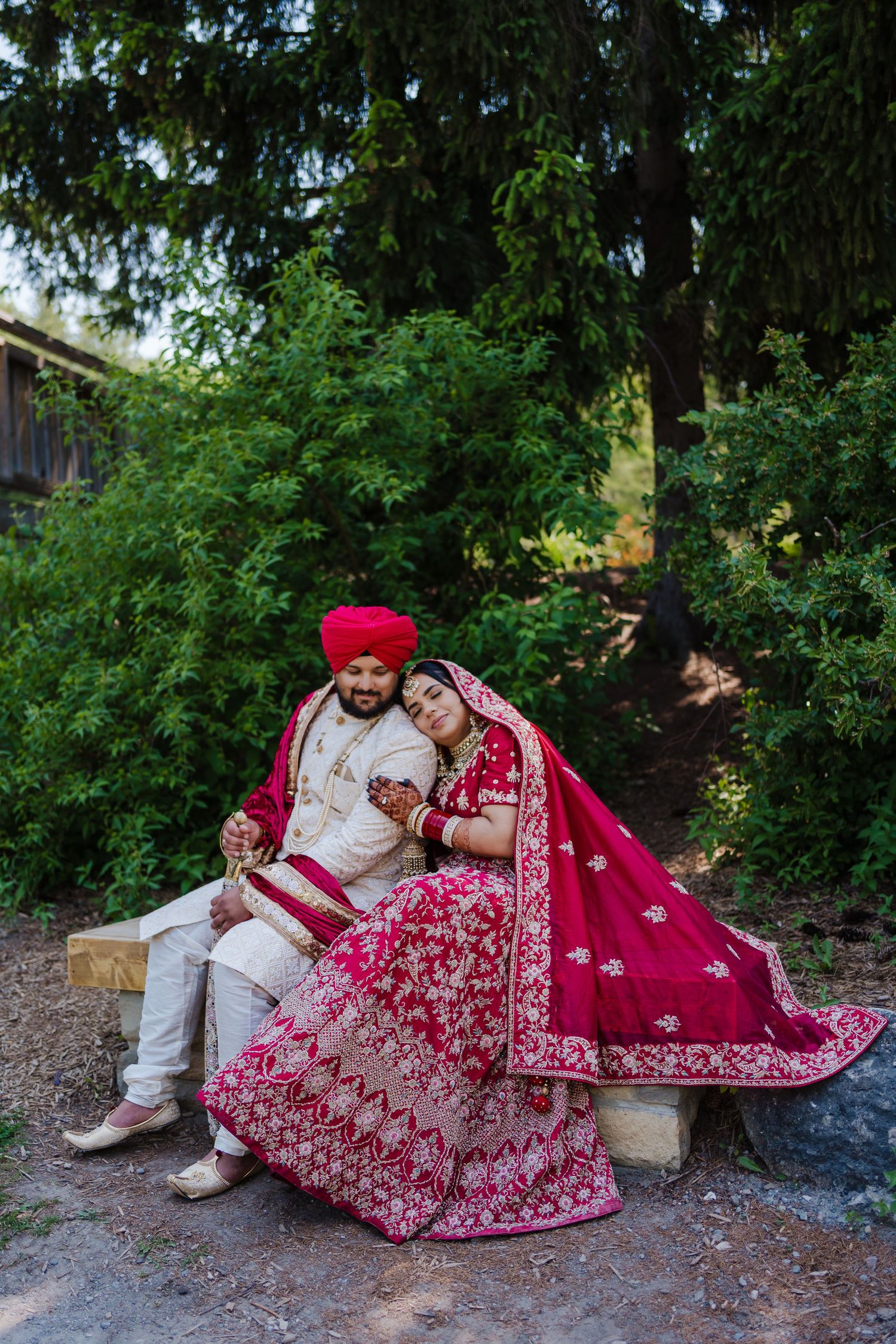 Traditional Sikh wedding couple in matching red and white outfits sit together outdoors among lush greenery.