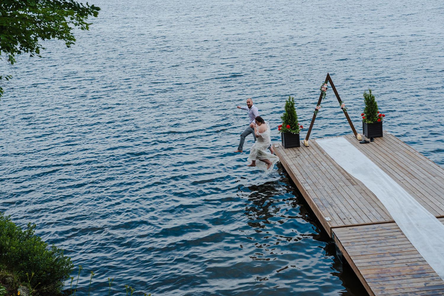 Person jumping off a wooden dock into calm blue lake water on a summer day with potted plants on the dock.