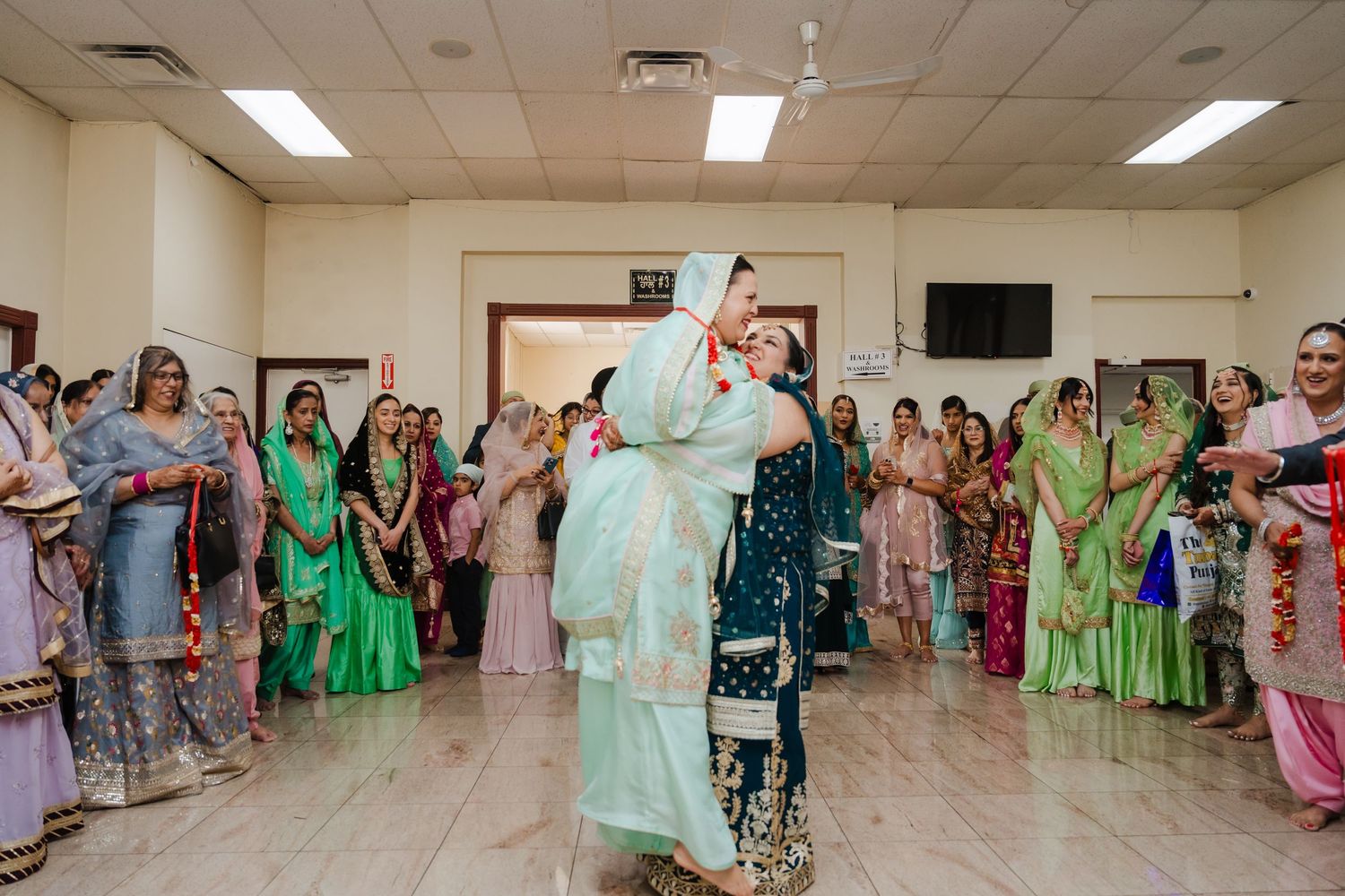 Wedding guests in traditional Indian attire form a circle during a celebratory dance in a community hall.