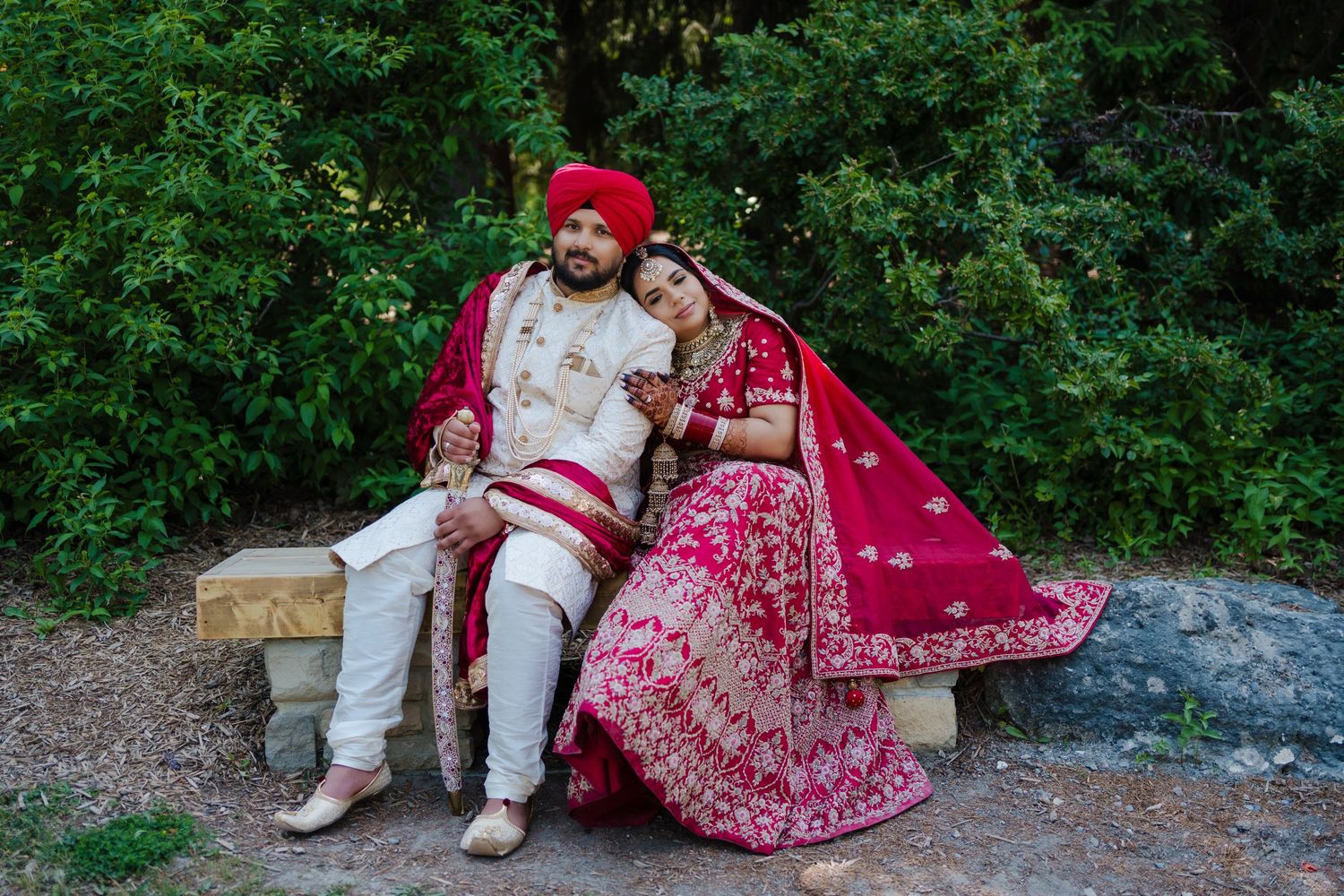 Traditional Indian wedding couple in red and white outfits sitting outdoors against green foliage.