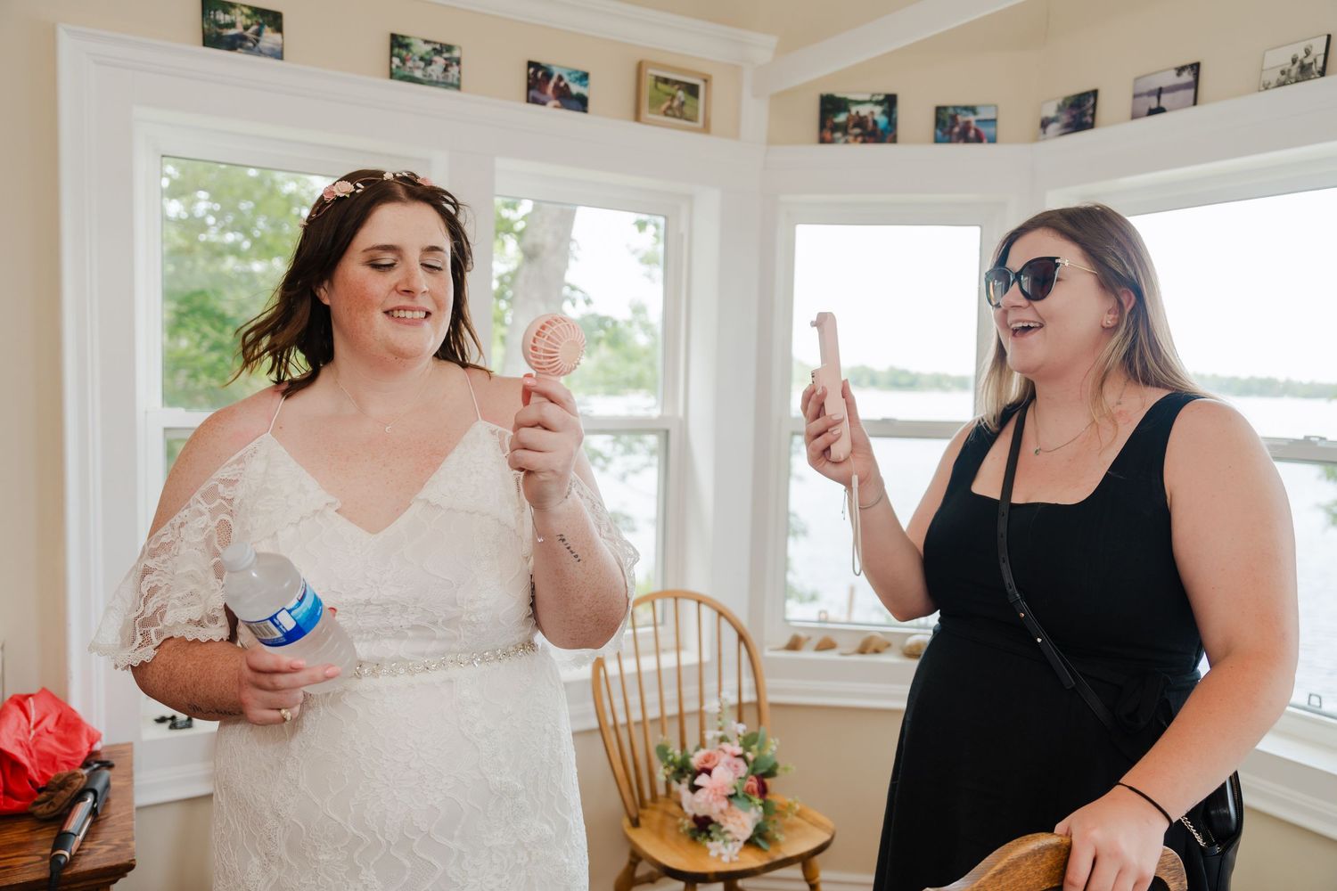 Two women share a joyful moment while getting ready in a bright sunlit room with photos on the wall.