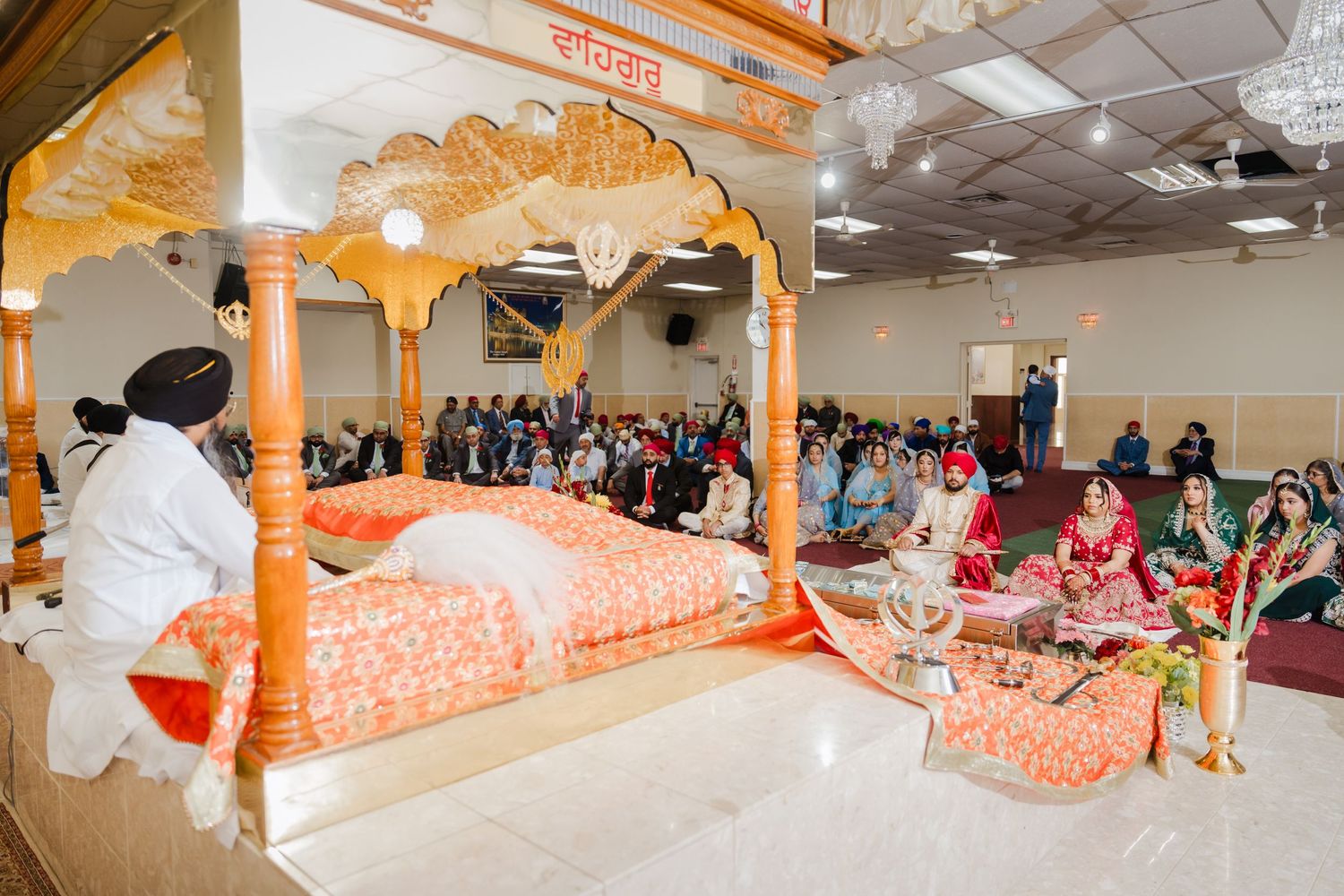 People gather in a Sikh temple gurdwara around an ornate orange and gold altar during a traditional religious ceremony.