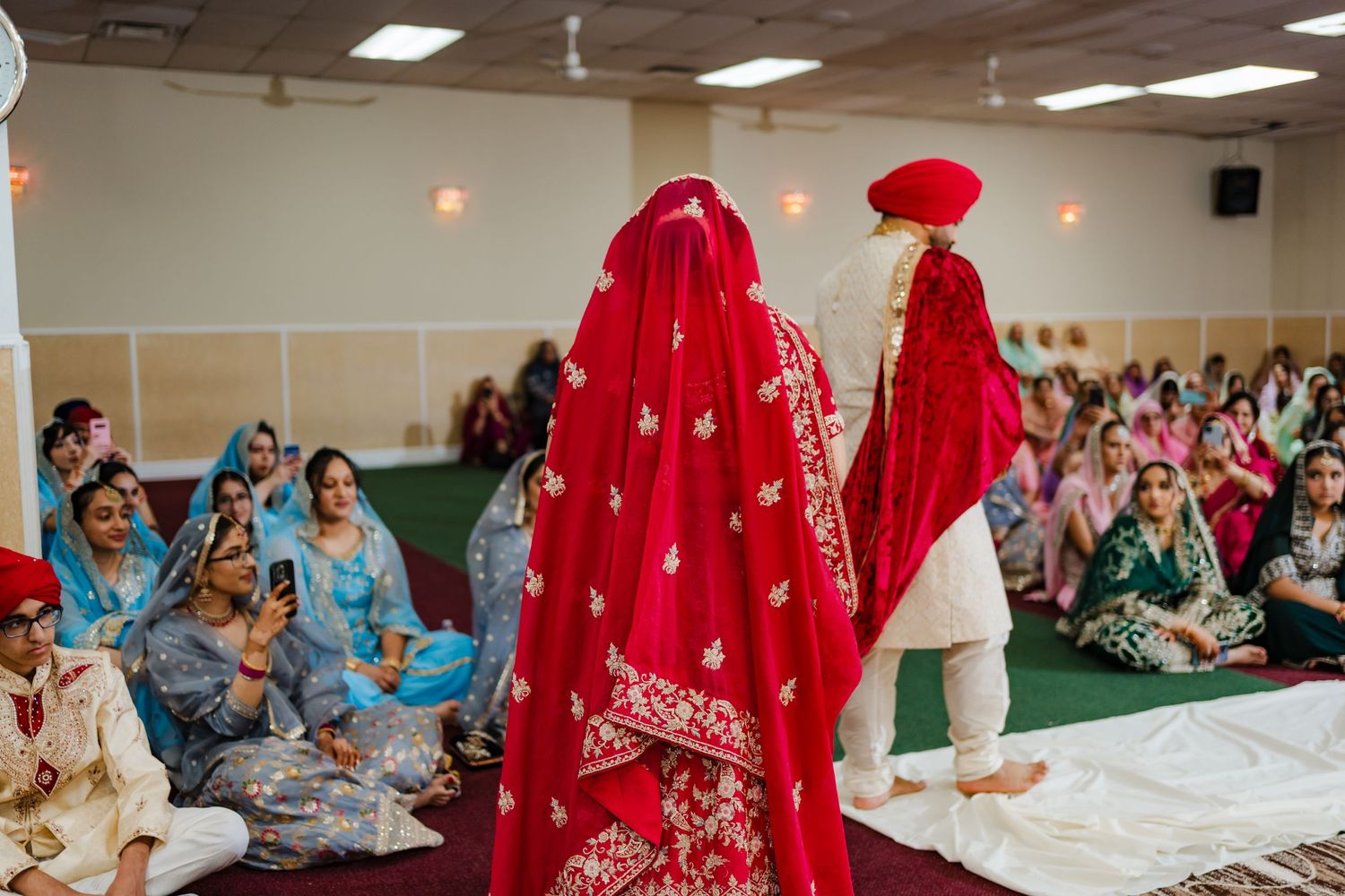 Sikh wedding ceremony with attendees in traditional attire seated on white floor coverings during indoor celebration.